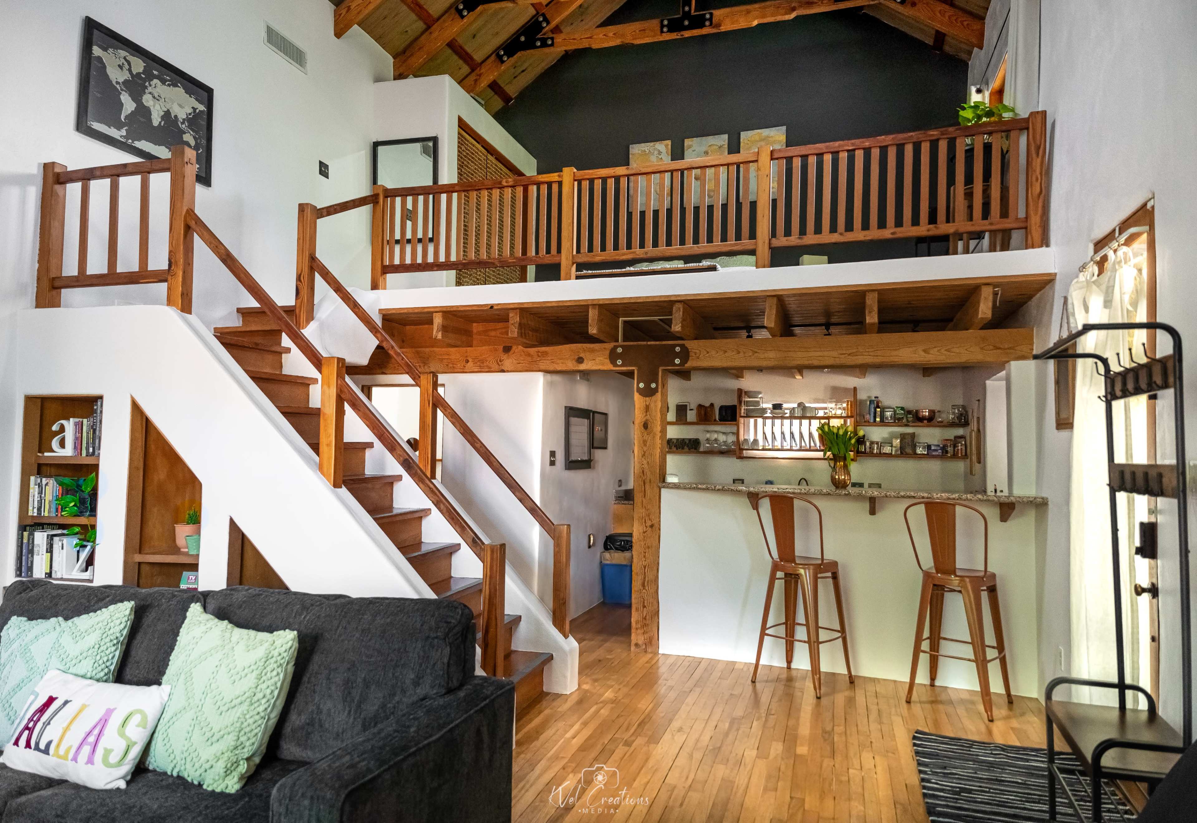 The image shows a spacious interior of a house with a loft, featuring a staircase, wooden beams, a seating area with a gray couch, and a bar with two stools in the background.