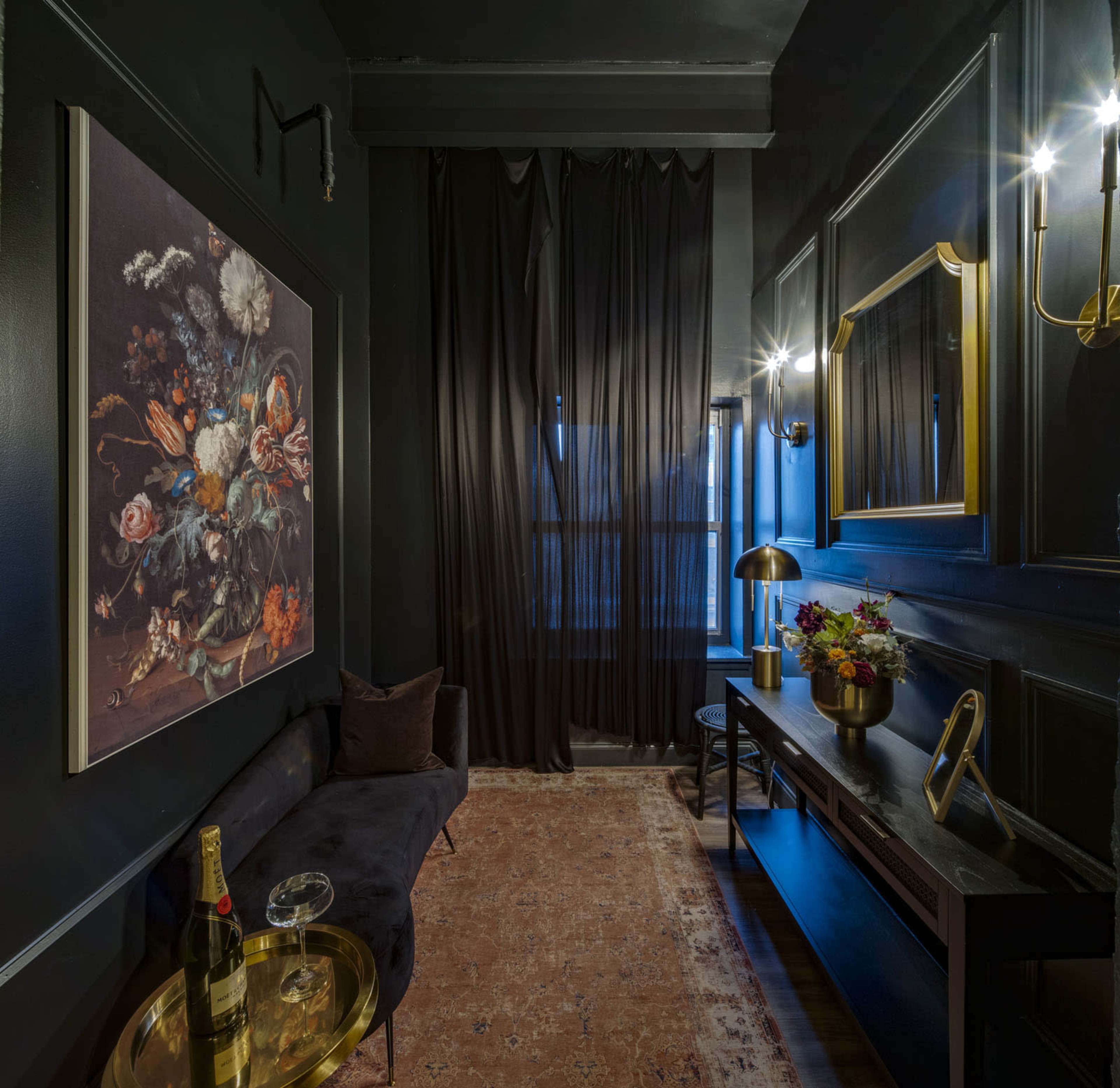 A dimly lit, elegantly furnished room featuring a dark color palette, a floral painting on the wall, a black sofa, and a wooden console table with decorative items and a bottle of champagne.