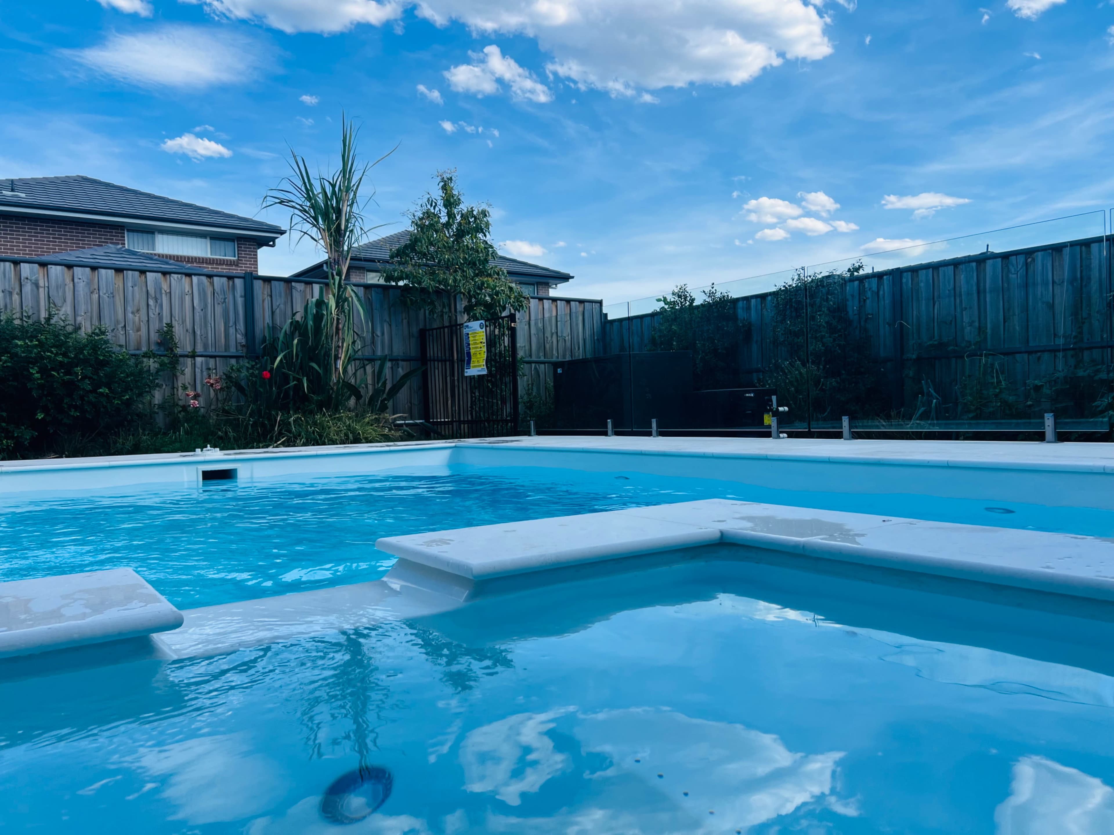 The image shows a clear blue swimming pool surrounded by a wooden fence and greenery under a partly cloudy sky.