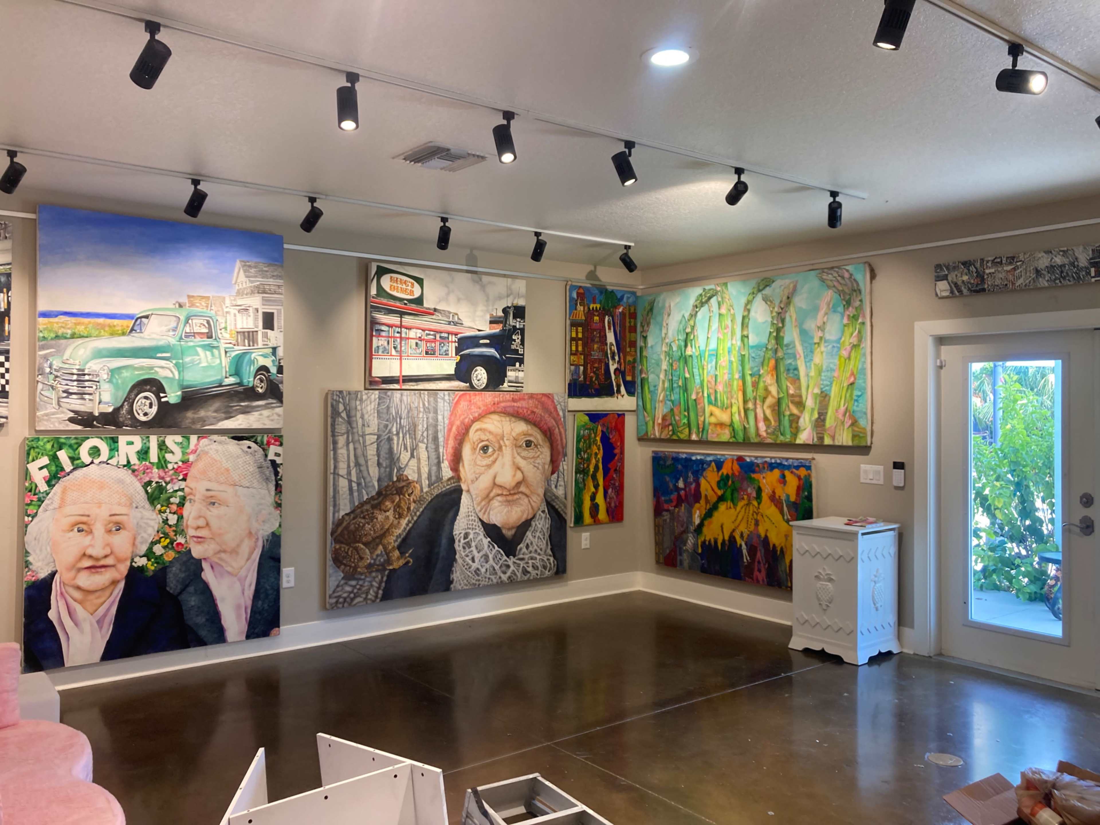 The image shows an art gallery with multiple colorful paintings displayed on the walls, featuring a variety of subjects including landscapes and portraits.