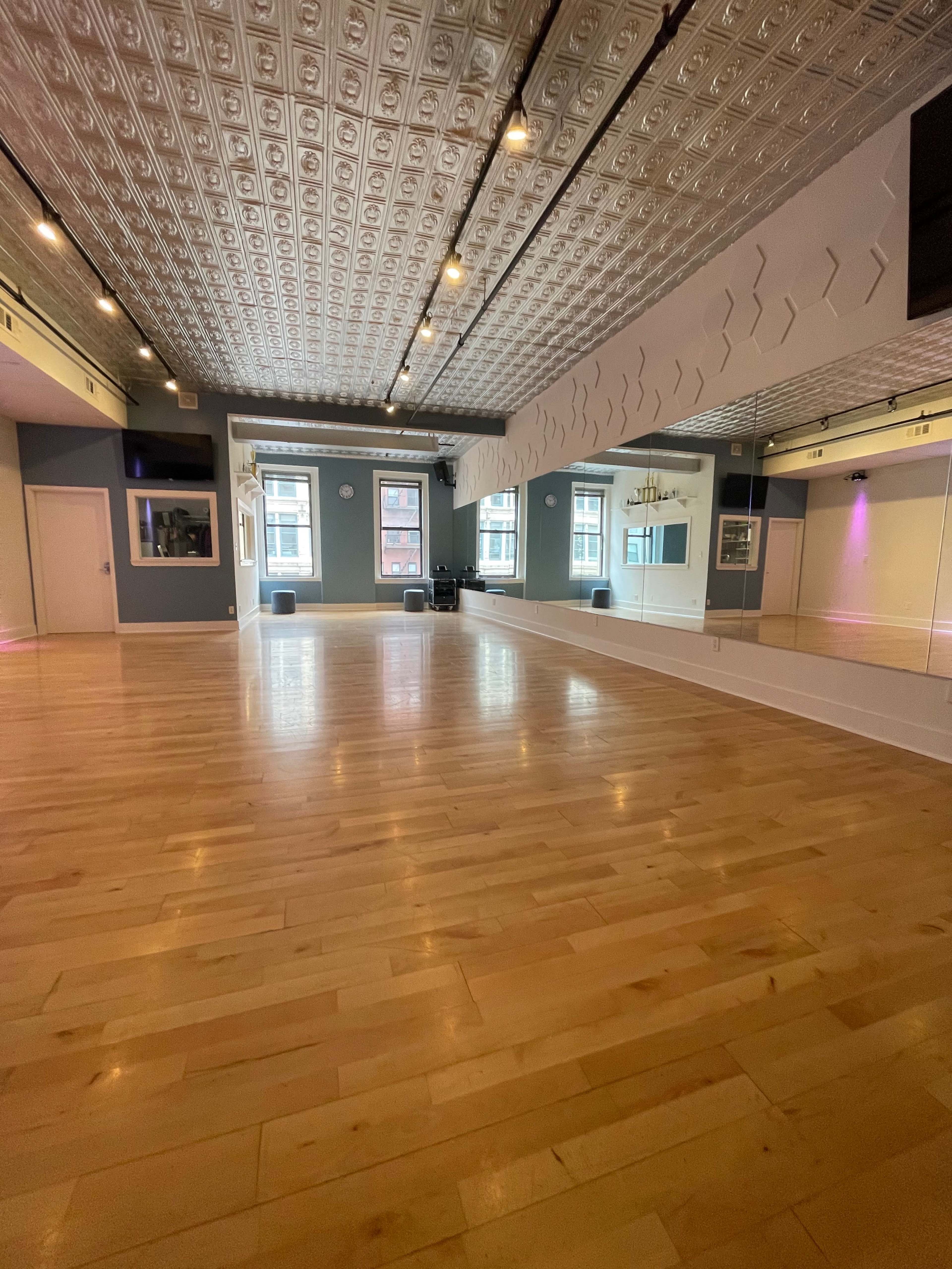 The image shows a spacious dance studio with wooden floors, mirrors, and decorative ceiling tiles, illuminated by soft lighting.