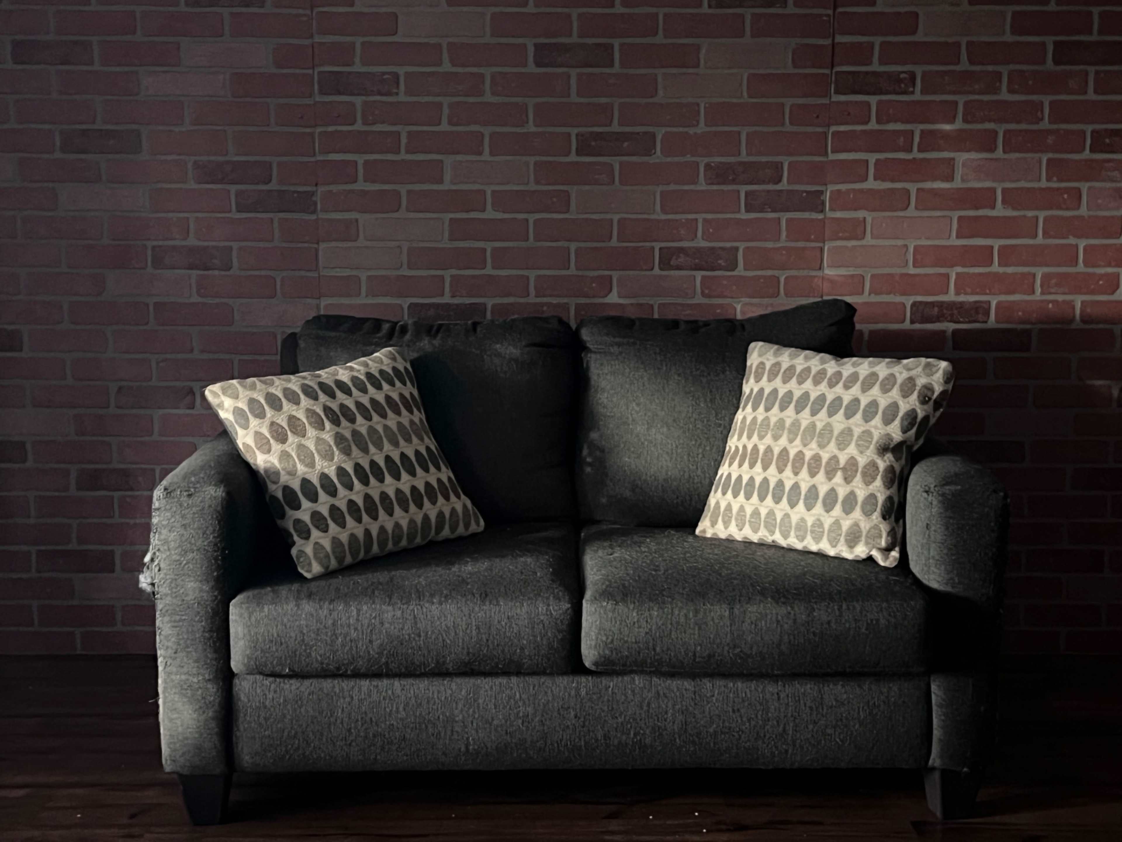 A dark gray loveseat with patterned cushions is positioned against a brick wall.