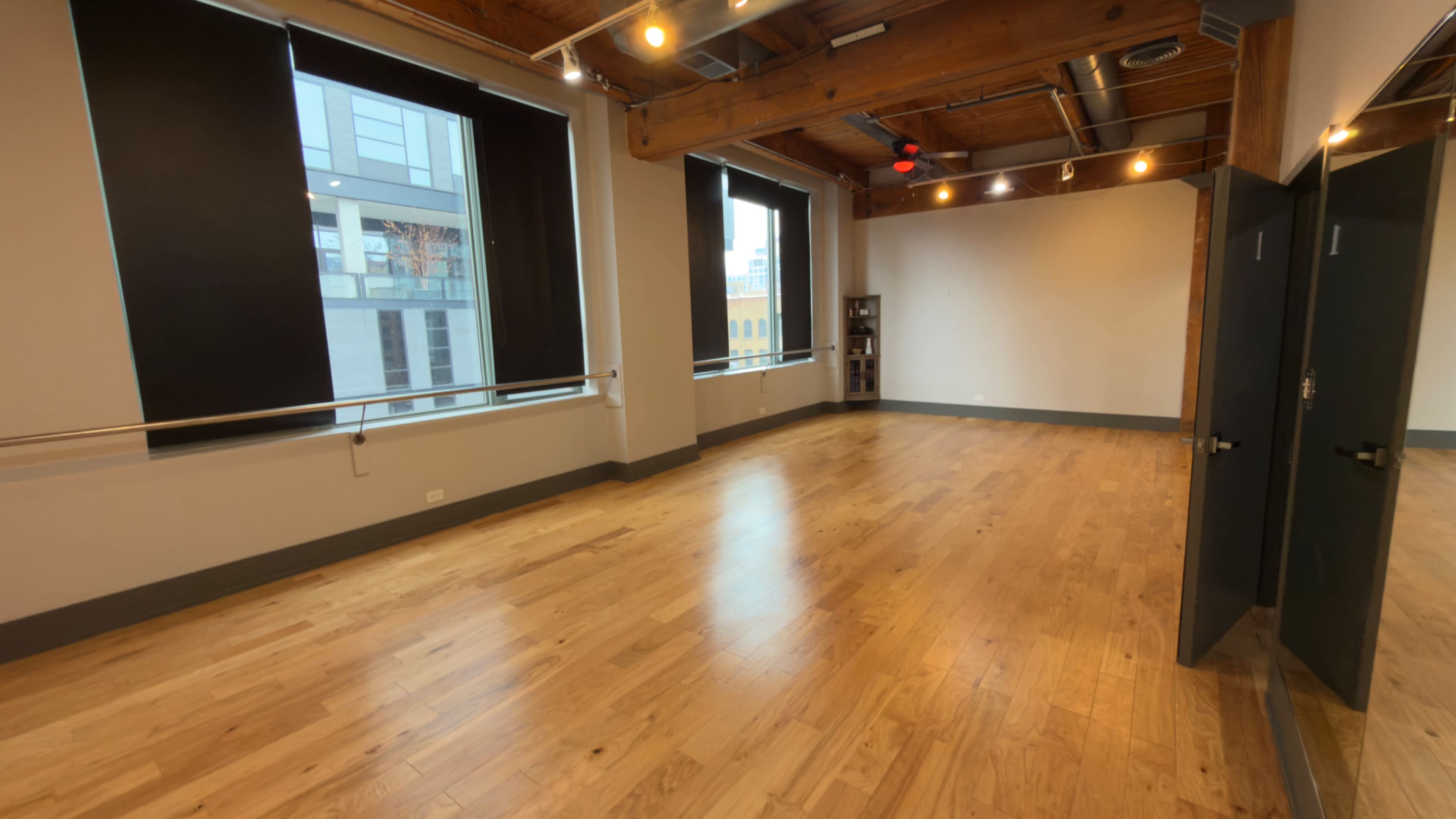 The image shows a spacious, empty room with hardwood floors, large windows, and exposed wooden beams.