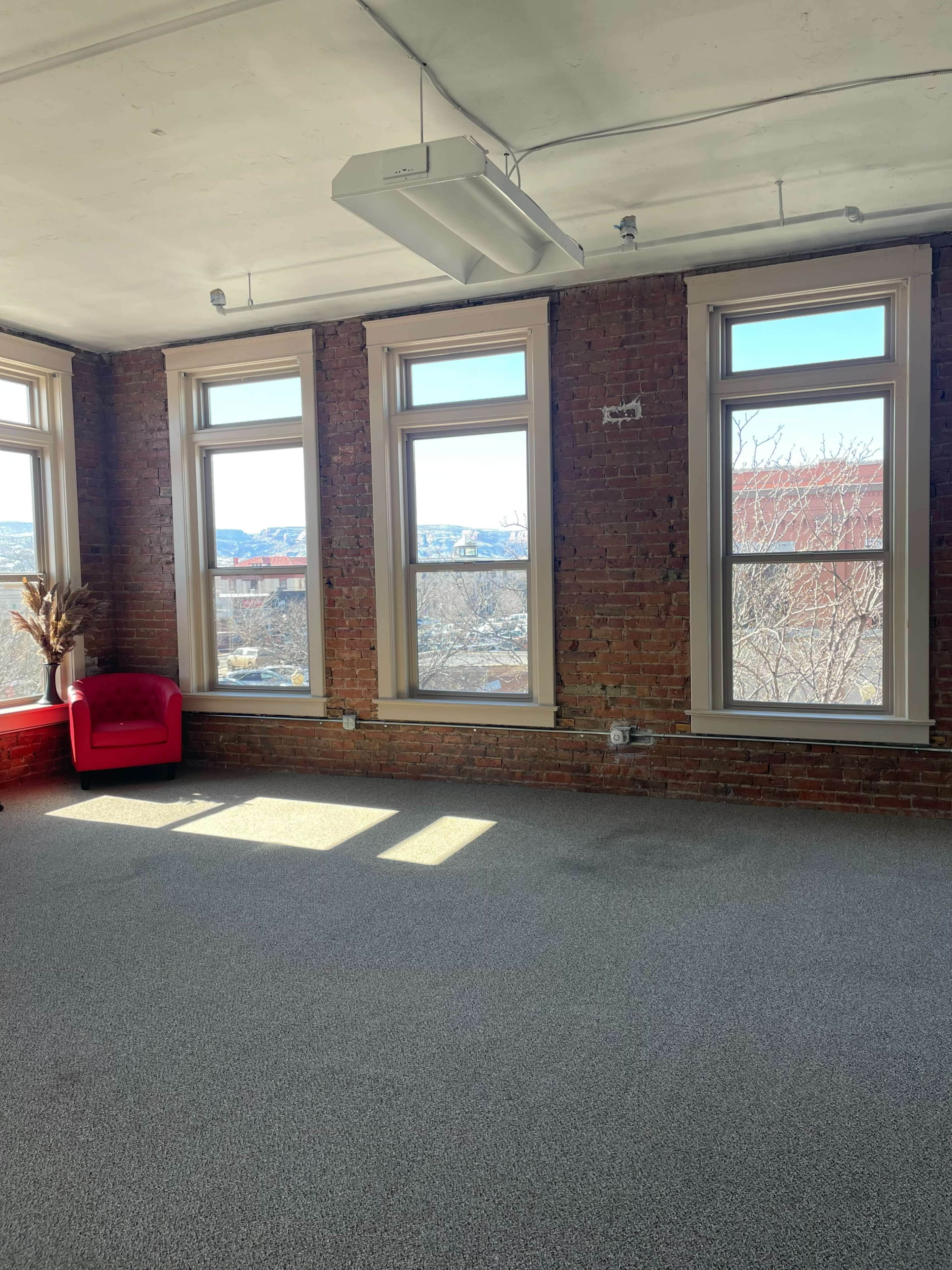 The image shows a spacious room with exposed brick walls, three large windows allowing natural light, and a single red chair in one corner.