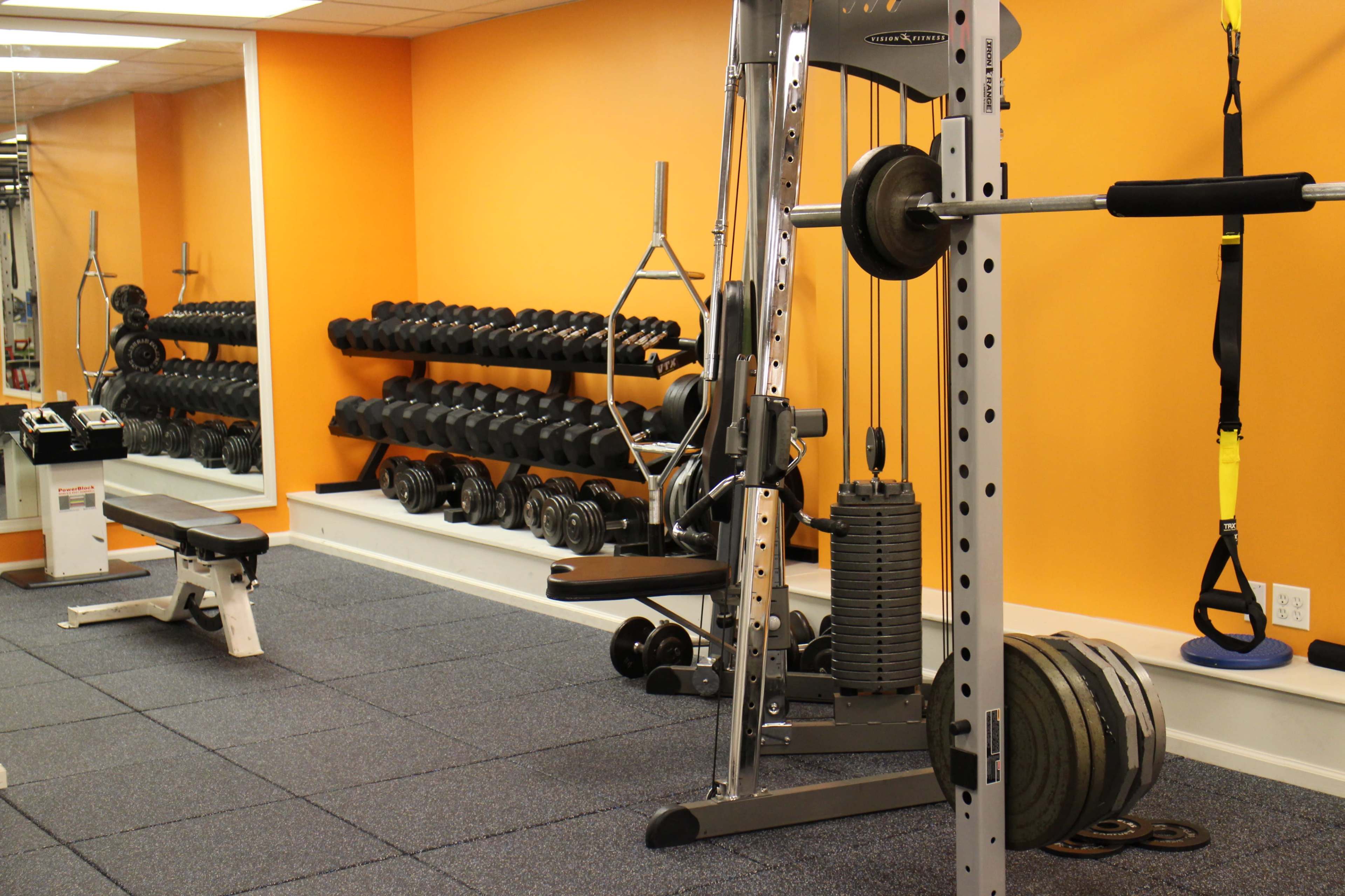 The image shows a gym interior with a strength training machine, a weight bench, and racks of dumbbells against an orange wall.