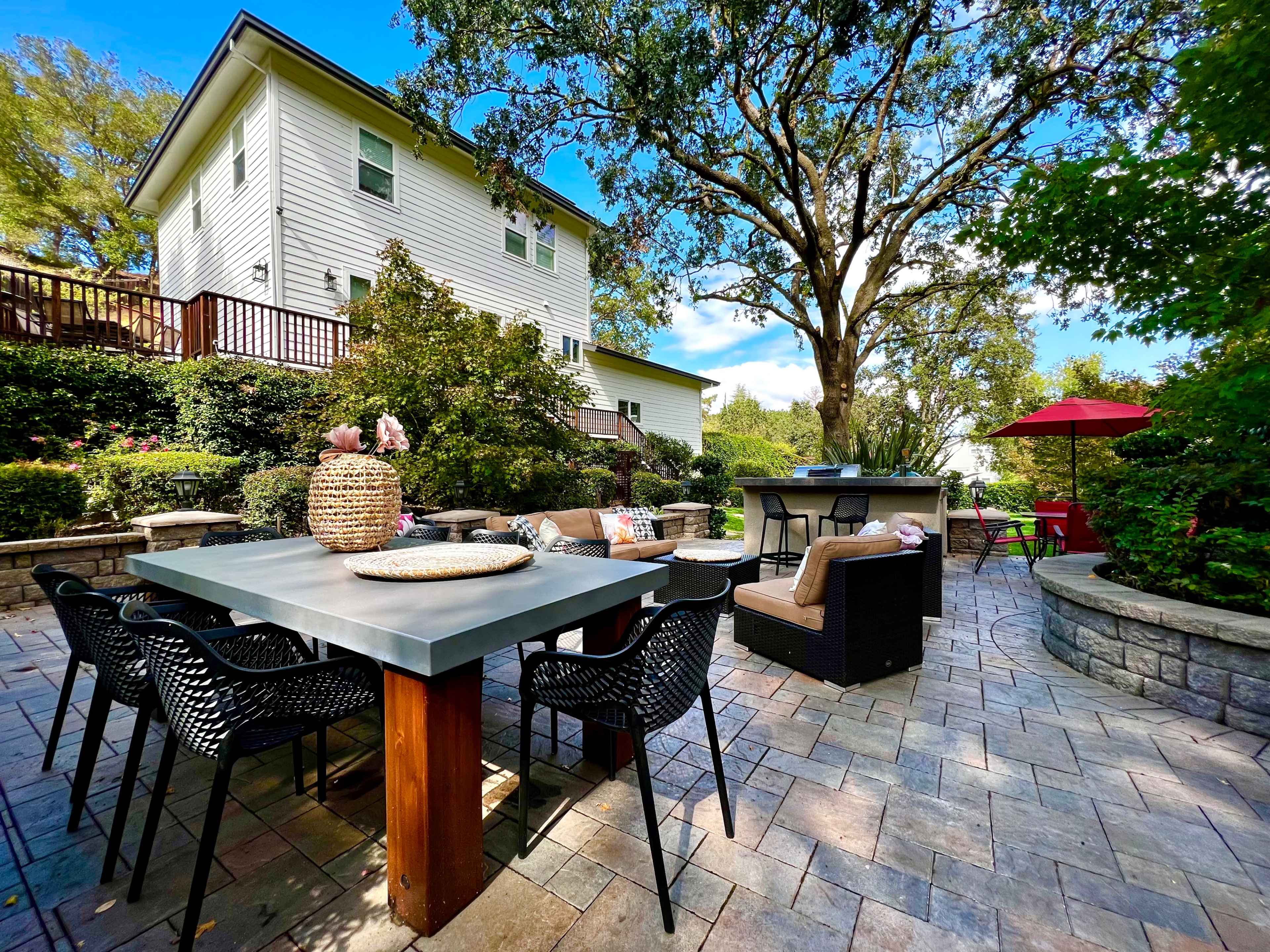 The scene shows a patio area with a large table, chairs, and a barbecue station, surrounded by greenery and a white house in the background.