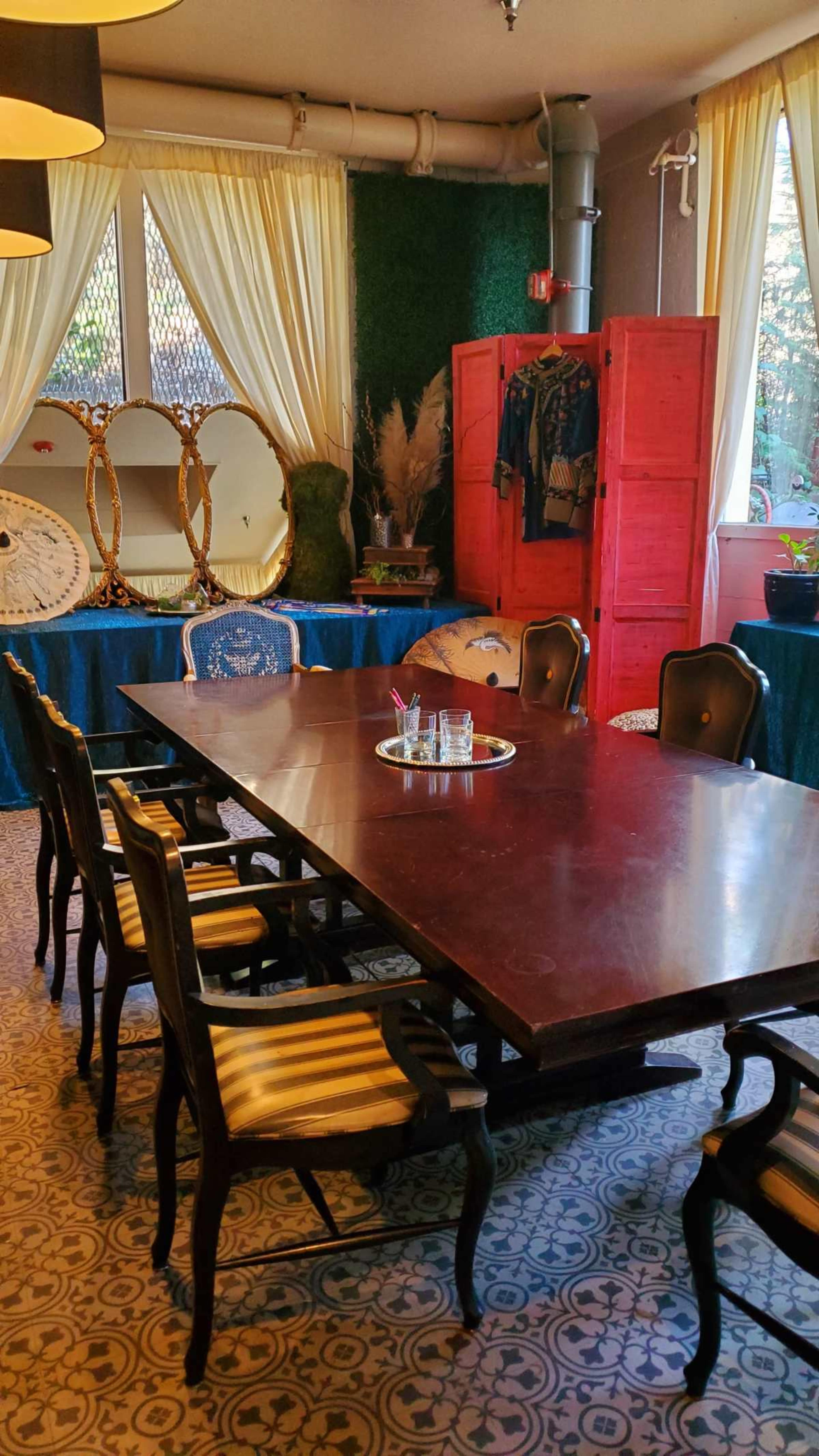A long wooden table is set in a room with large windows, surrounded by dark chairs, with a red cabinet and decorative mirrors in the background.