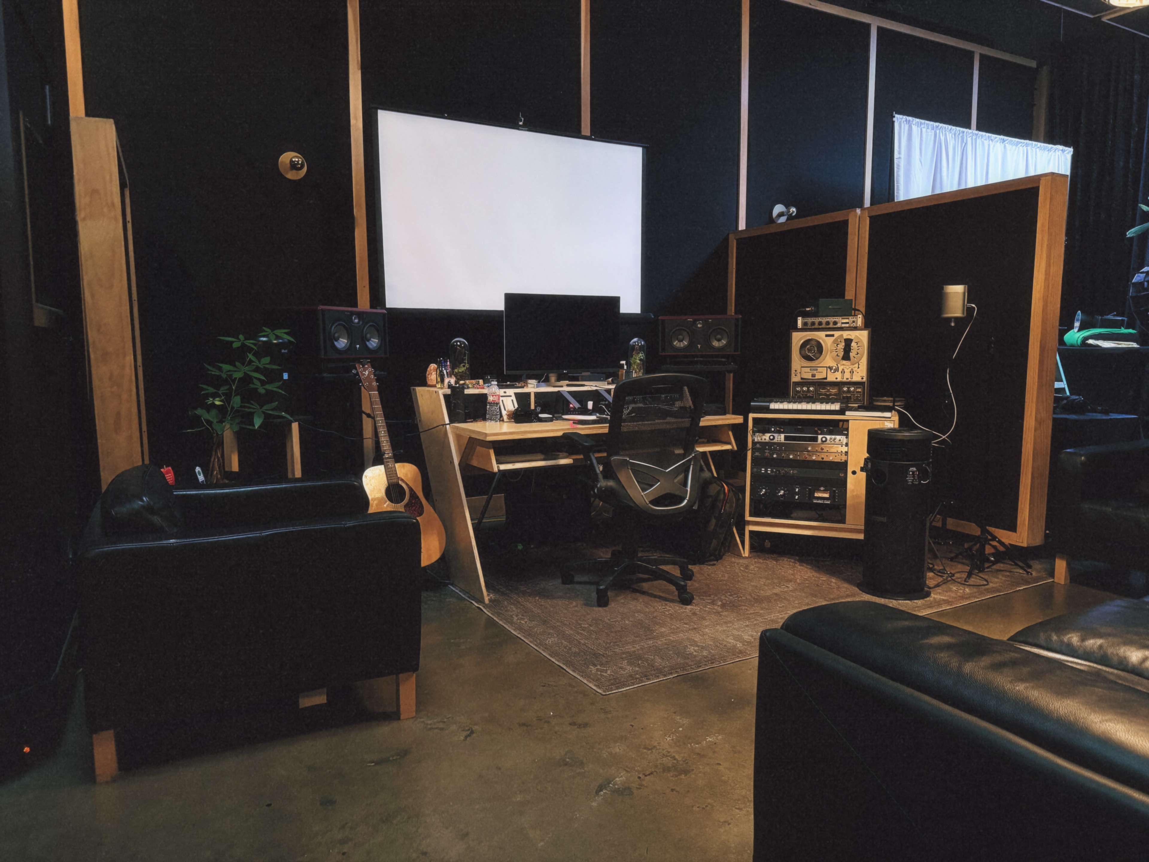 The image shows a music studio featuring a desk with a computer, audio equipment, comfortable chairs, and plants, all within a dimly lit space.