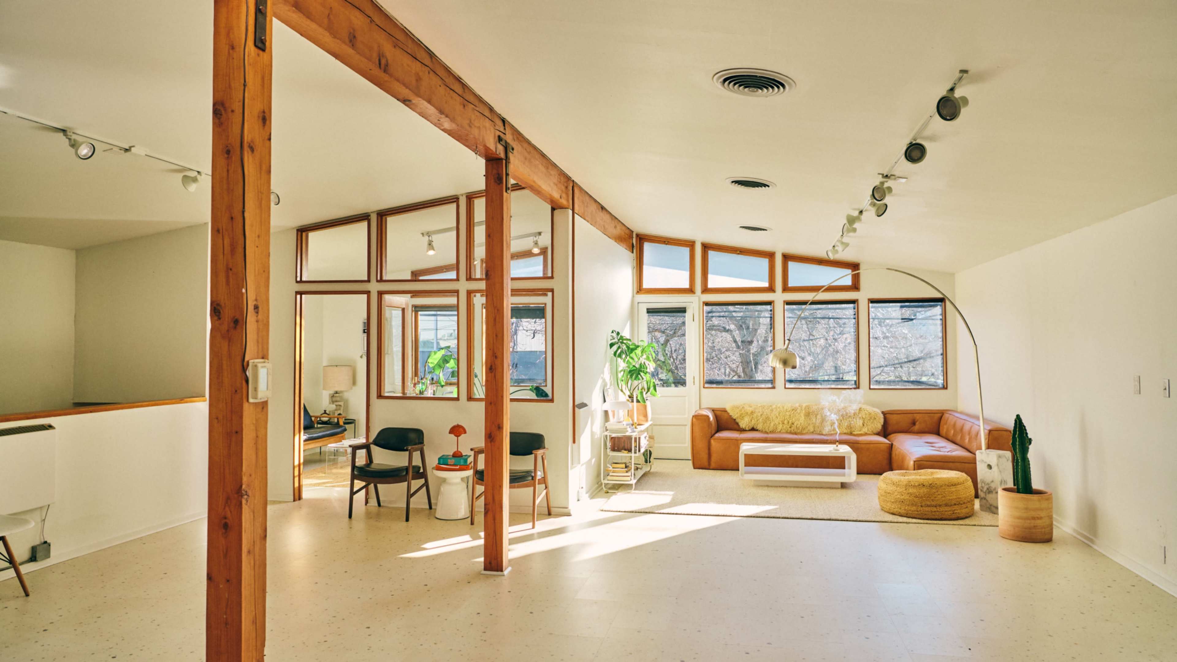 The image shows a spacious living area with large windows, wooden beams, and modern furniture, including a brown leather sofa and circular rug.