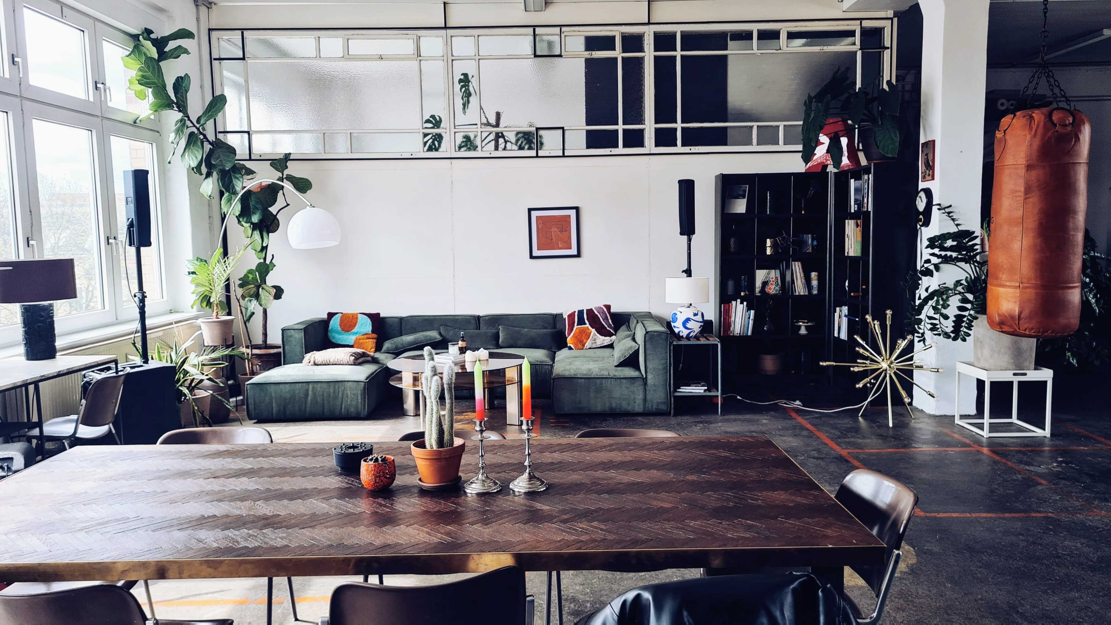 The image shows a modern, open living space with a green sofa, a wooden dining table, various plants, and a punching bag against the wall.