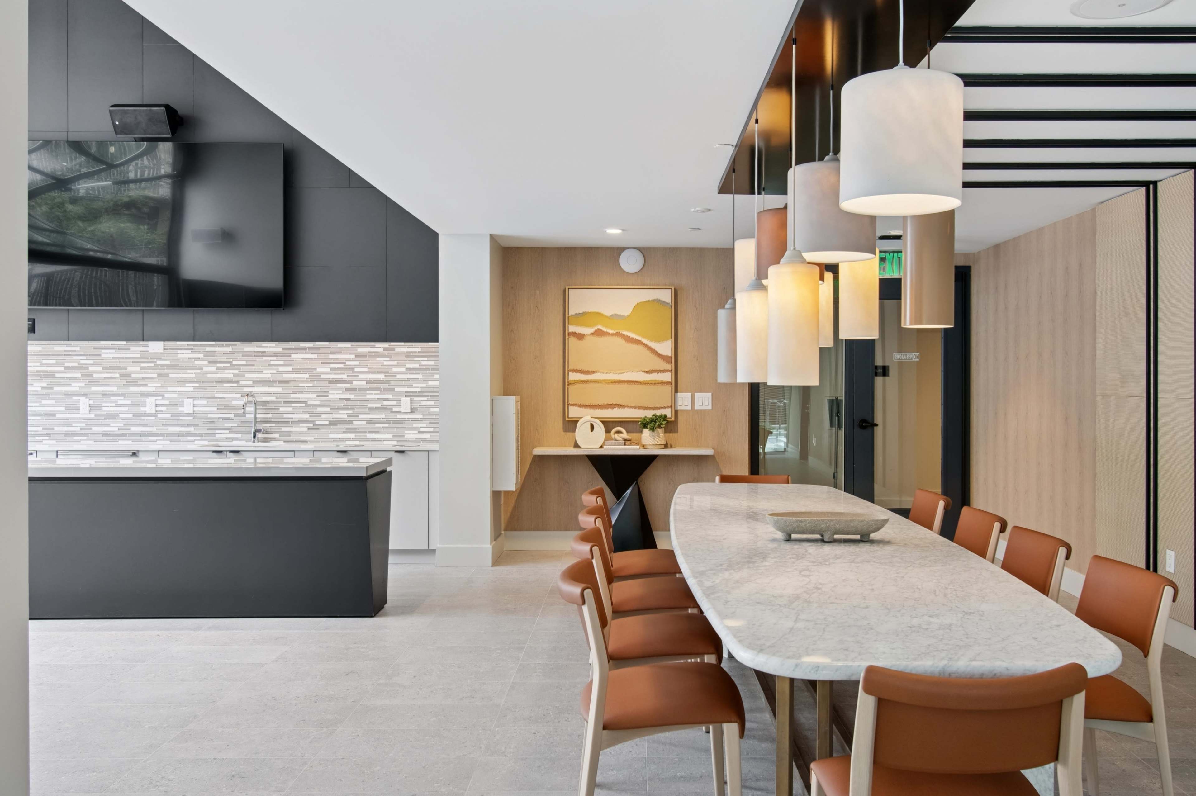 A modern kitchen and dining area features a long marble table surrounded by brown chairs, with a sleek black counter, pendant lighting, and a television on the wall.