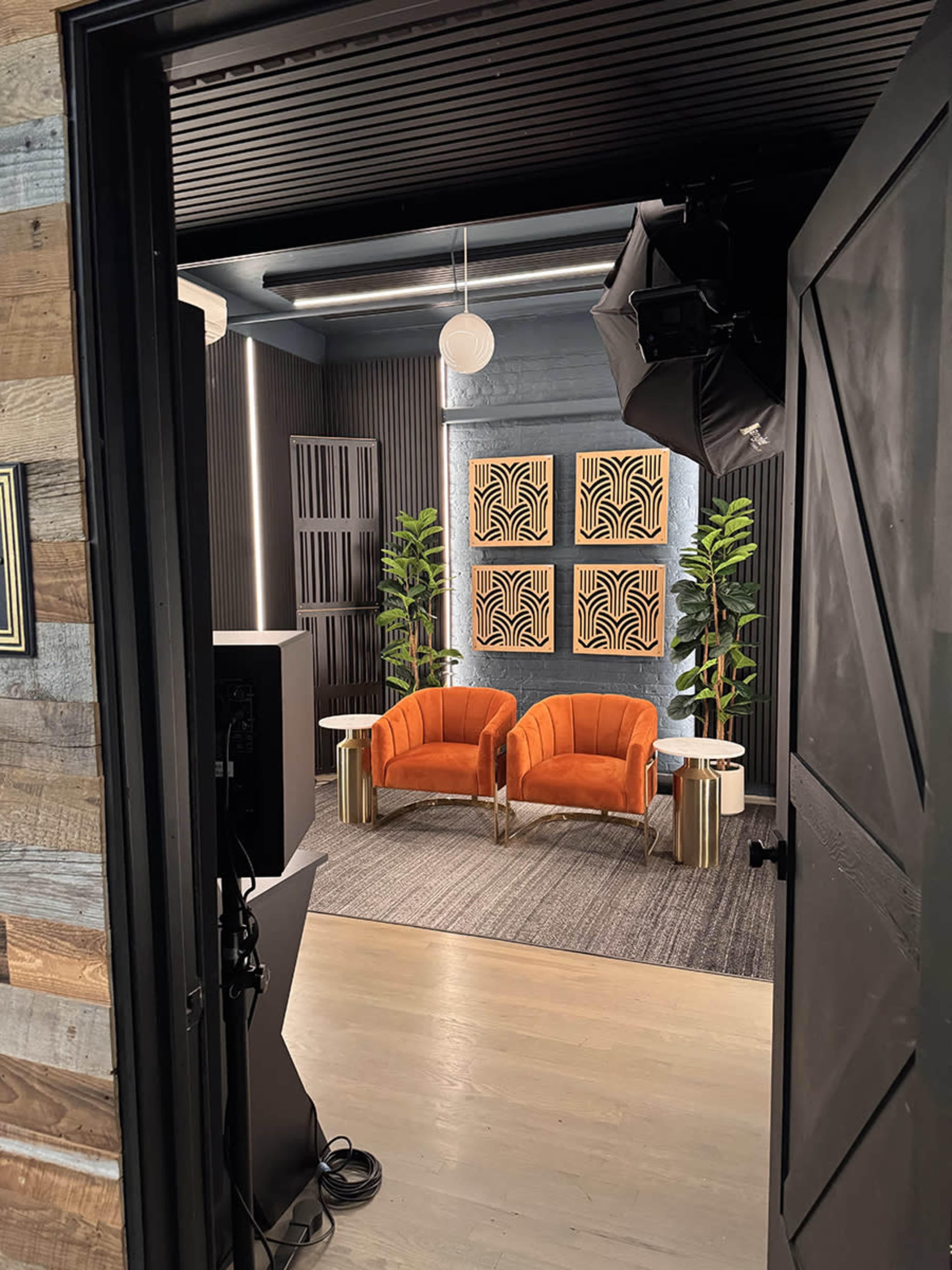 The image shows a modern interior with two orange velvet chairs positioned in front of decorative wall panels and potted plants, viewed through an open door.