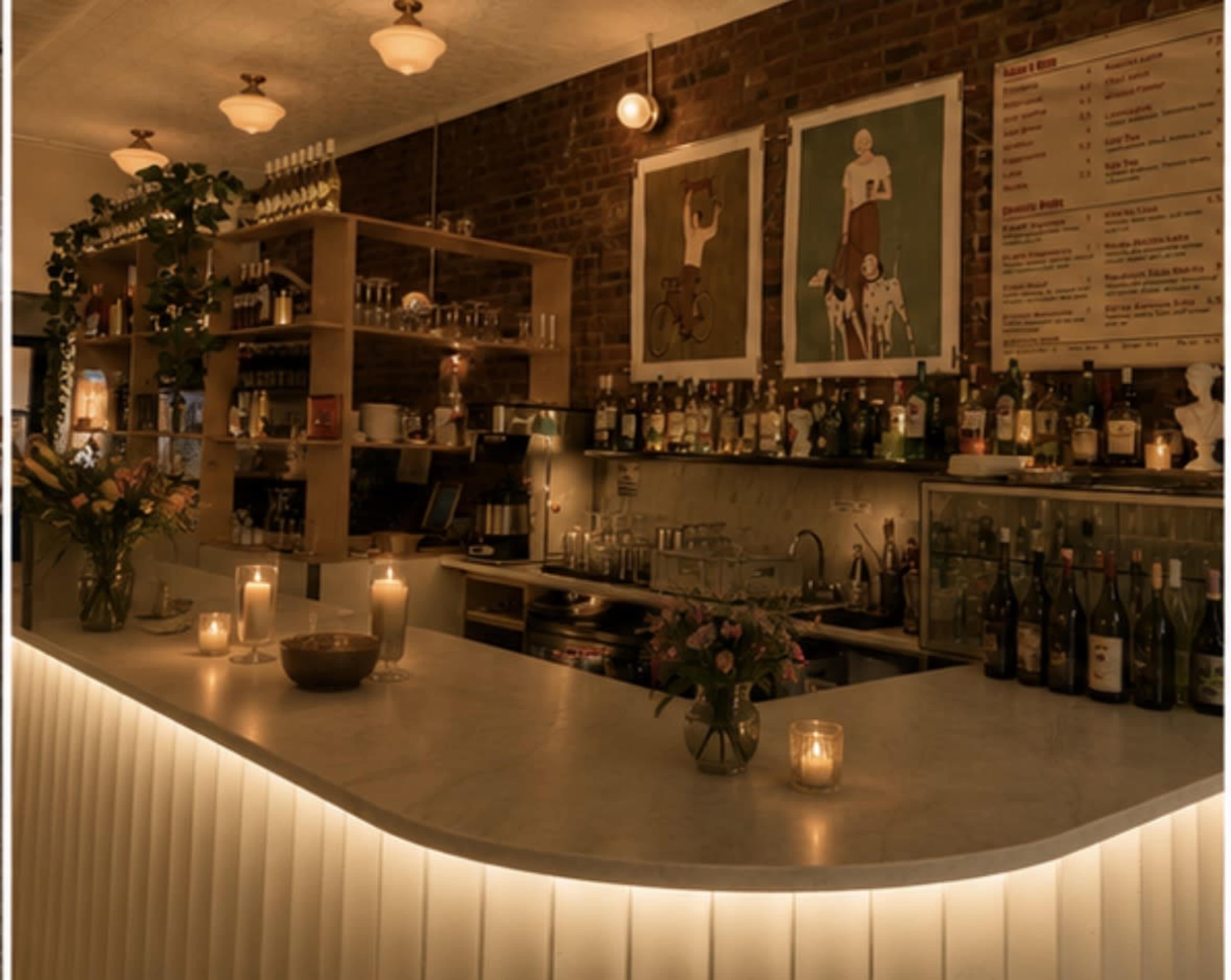 The image shows a well-lit bar area with a curved counter, shelves filled with various bottles, and decorative plants, set against a backdrop of exposed brick walls and artwork.