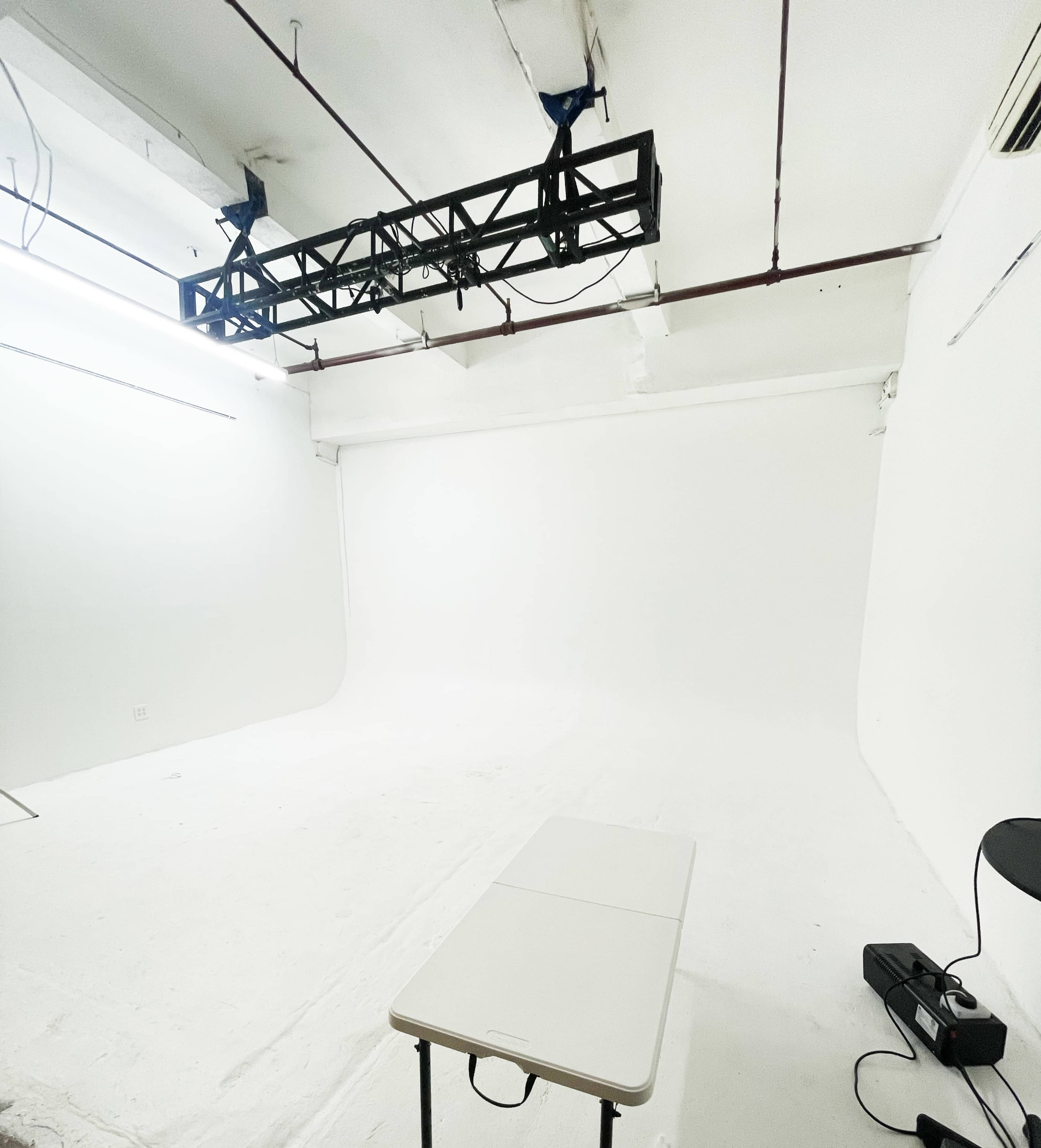 The image shows an empty studio space with white walls and a minimalistic setup, including a table in the foreground and a lighting rig suspended from the ceiling.