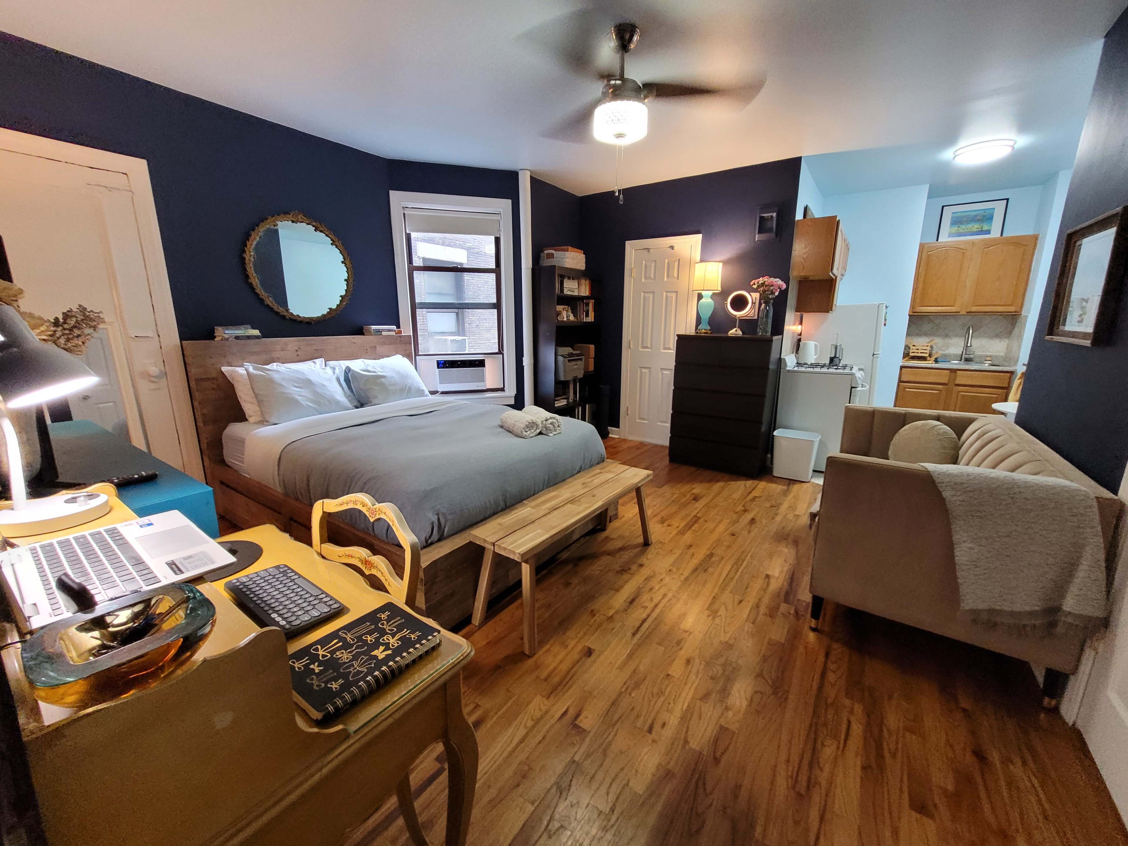 The image shows a cozy studio apartment featuring a bed, a small kitchenette, a seating area, and a desk with a computer.