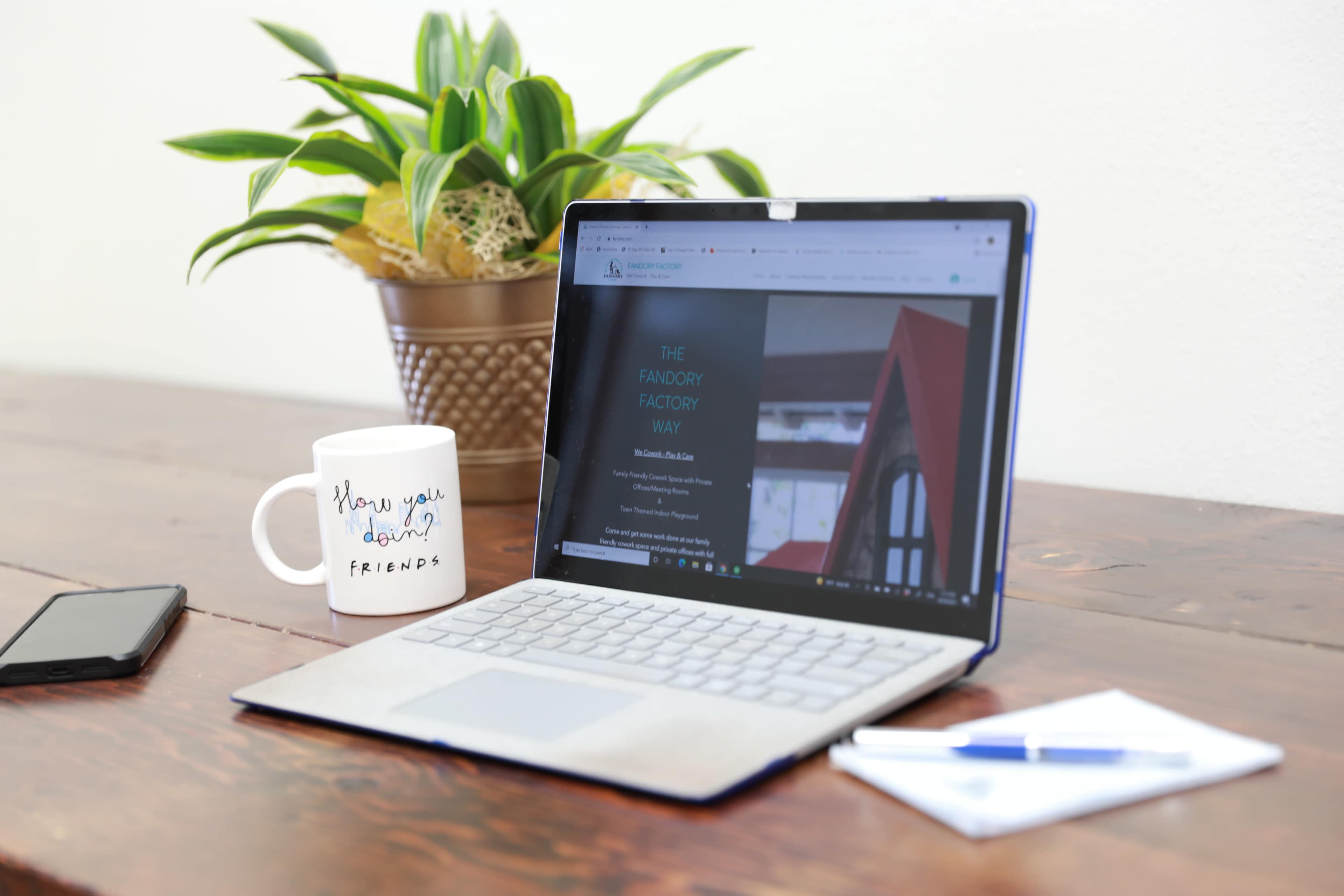 A silver laptop is open on a wooden table, displaying a webpage, next to a coffee mug with a greeting, a smartphone, a pen, and a notepad.