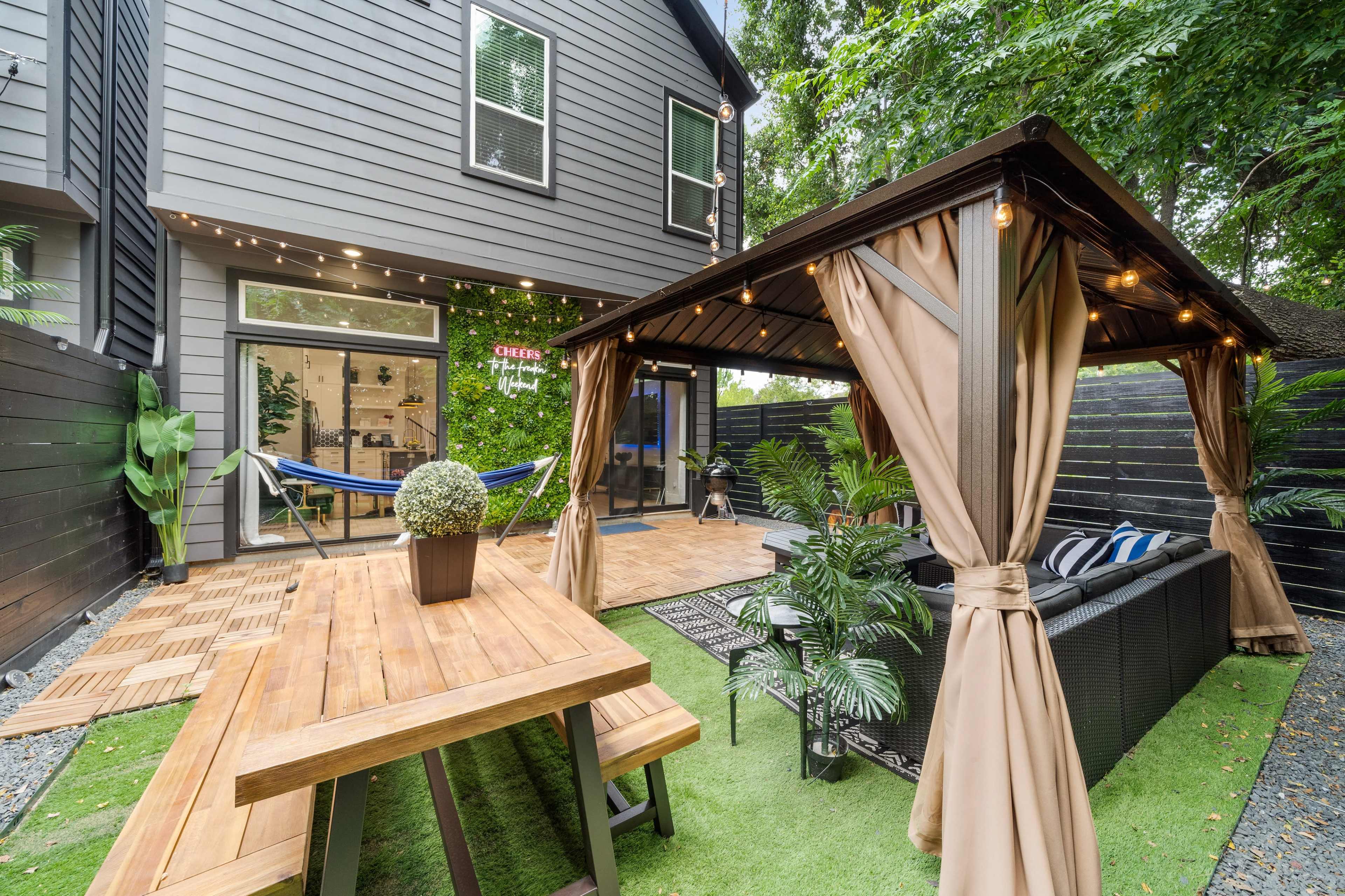 A landscaped backyard featuring a wooden dining table, a covered seating area with curtains, and decorative plants surrounded by a modern home.