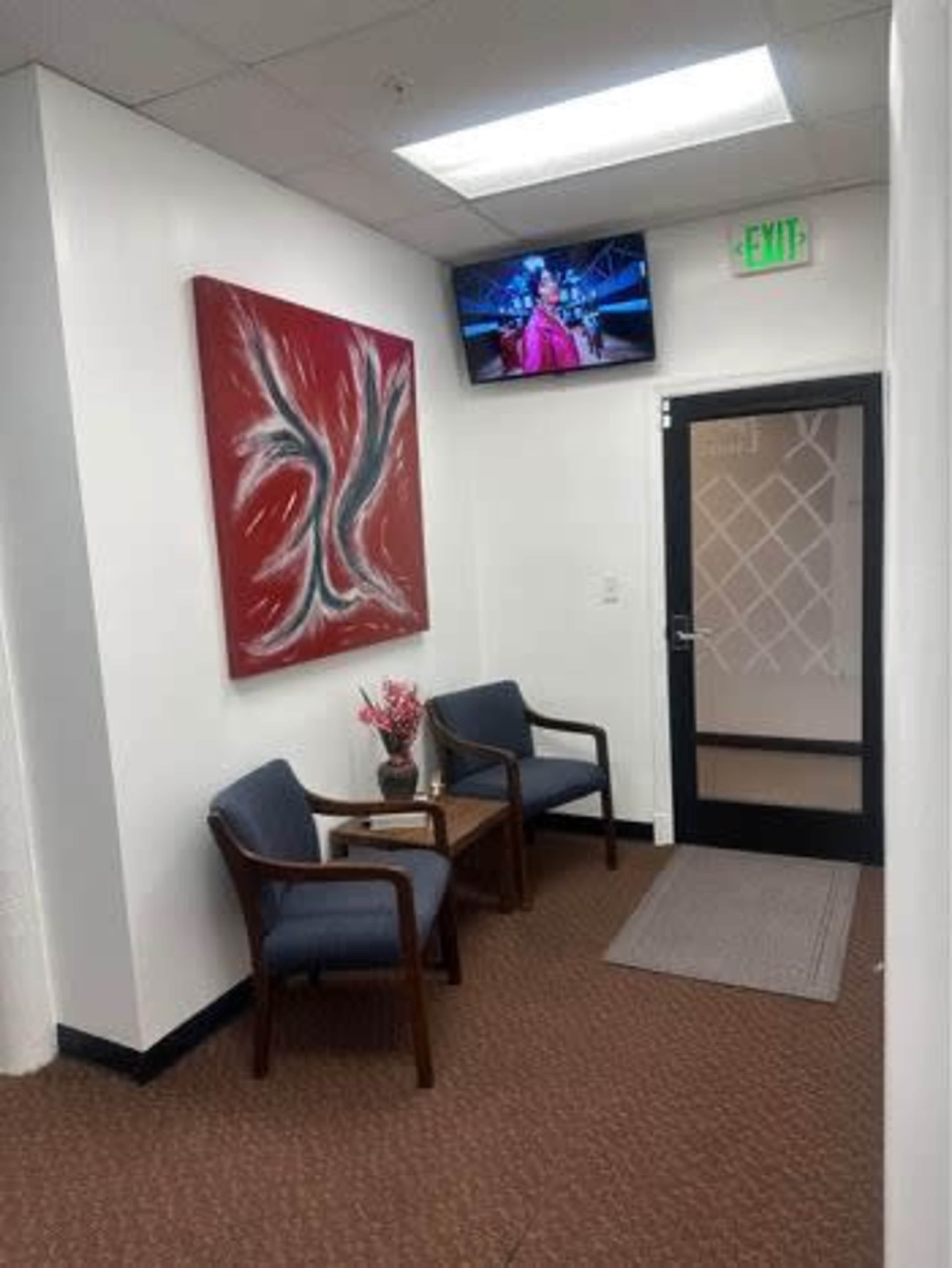 A small waiting area with two chairs, a side table, a piece of abstract art on the wall, and a TV mounted above displaying a program.