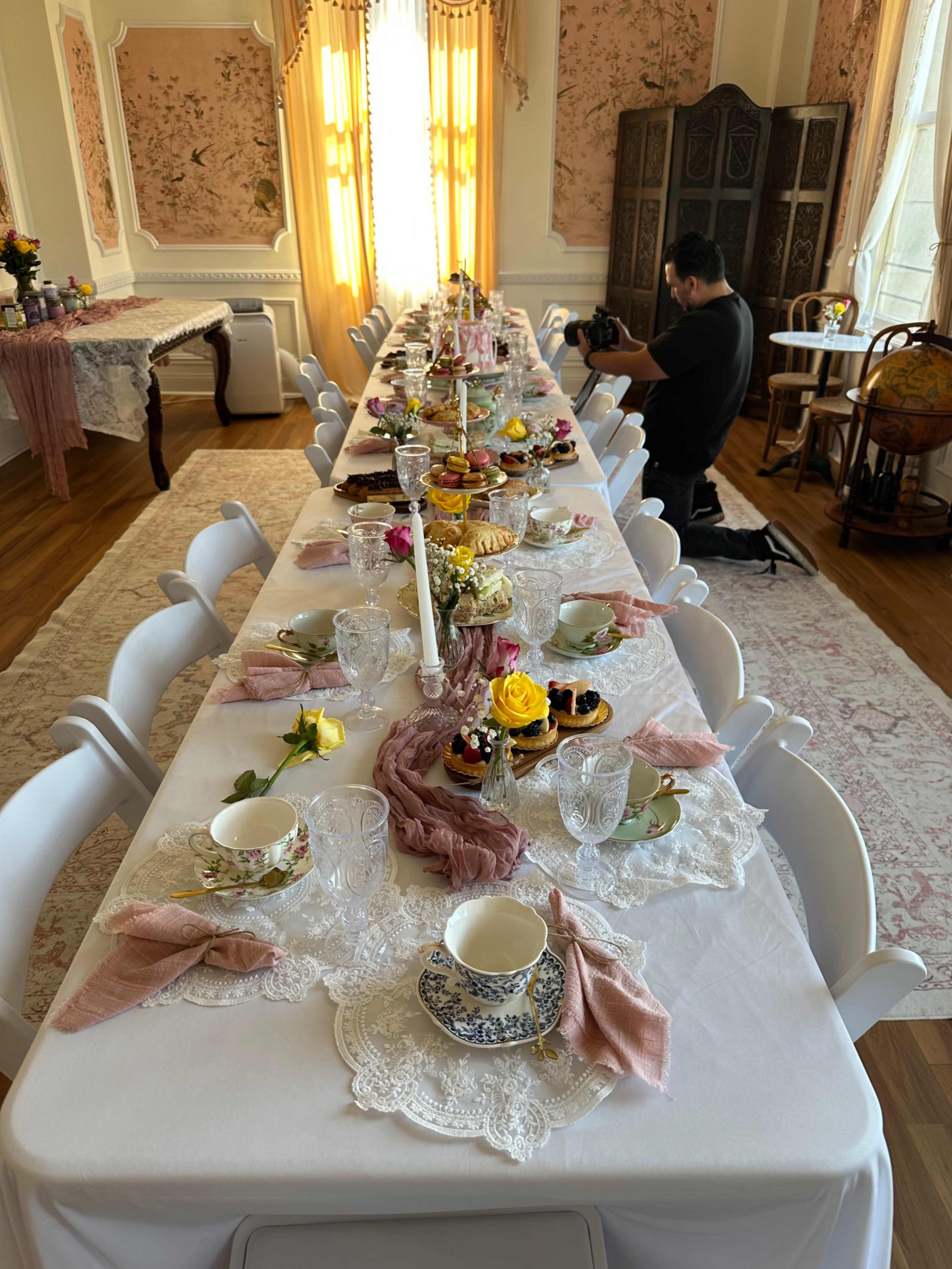 A long elegantly set dining table filled with various pastries, tea cups, and floral arrangements, with a bright window illuminating the scene.
