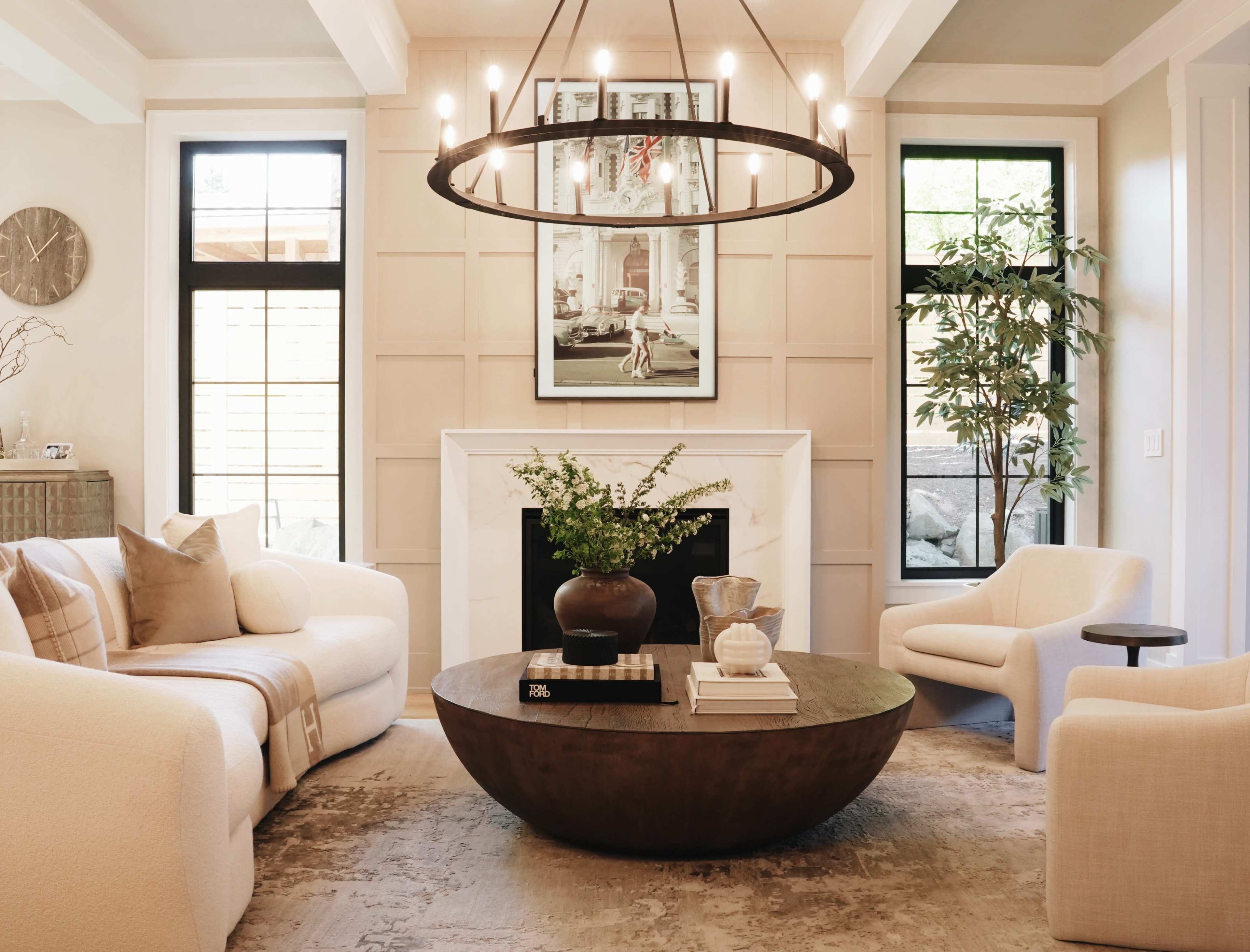 The image depicts a modern living room featuring a round wooden coffee table, a large overhead chandelier, and a cozy seating arrangement with light-colored armchairs and a sofa.