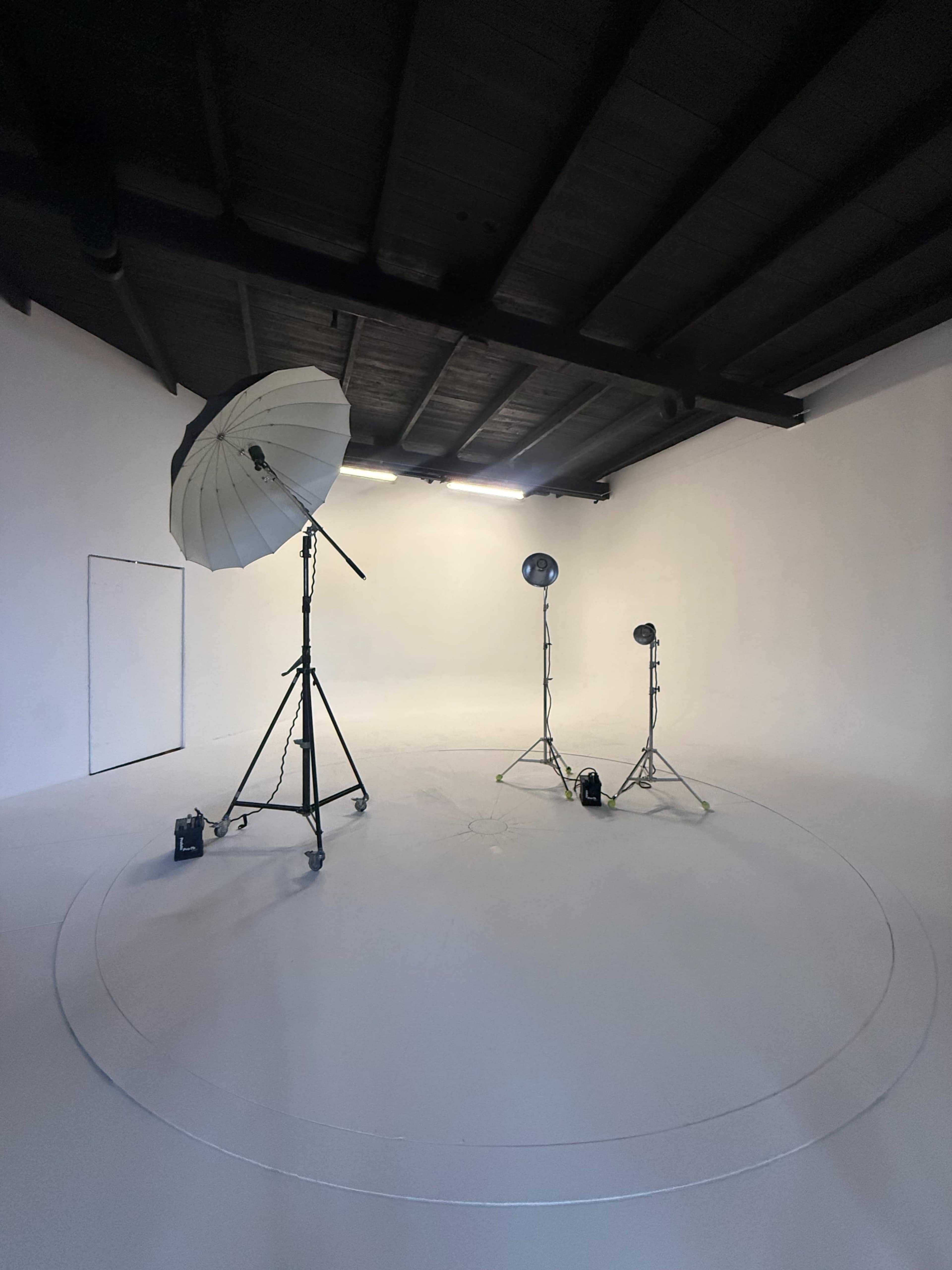 The image shows a minimalistic photo studio with a white backdrop, overhead lighting, and two light stands with an umbrella reflector.