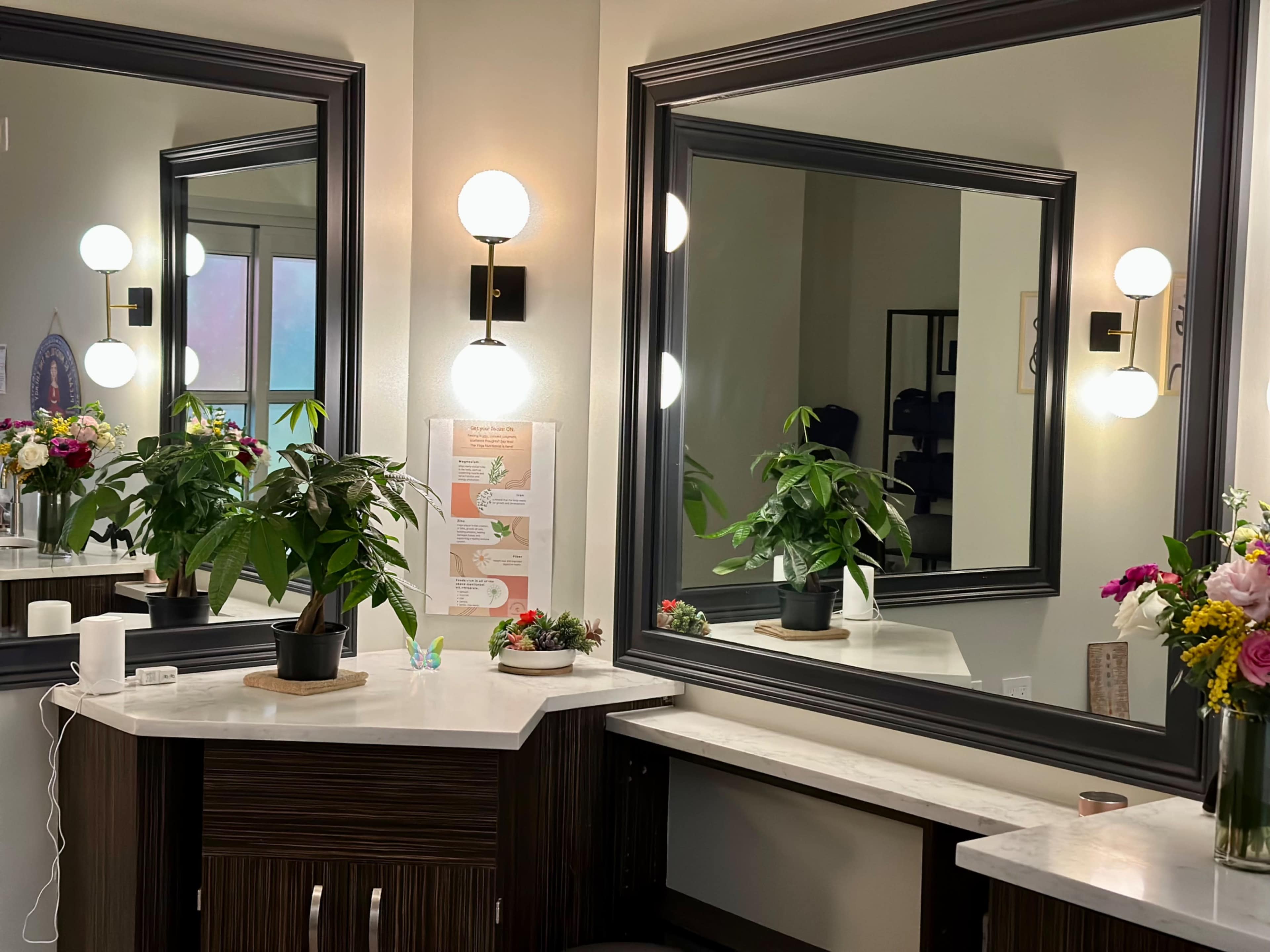The image shows a well-lit vanity area featuring a large mirror, decorative plants, and a bouquet of flowers on a countertop.