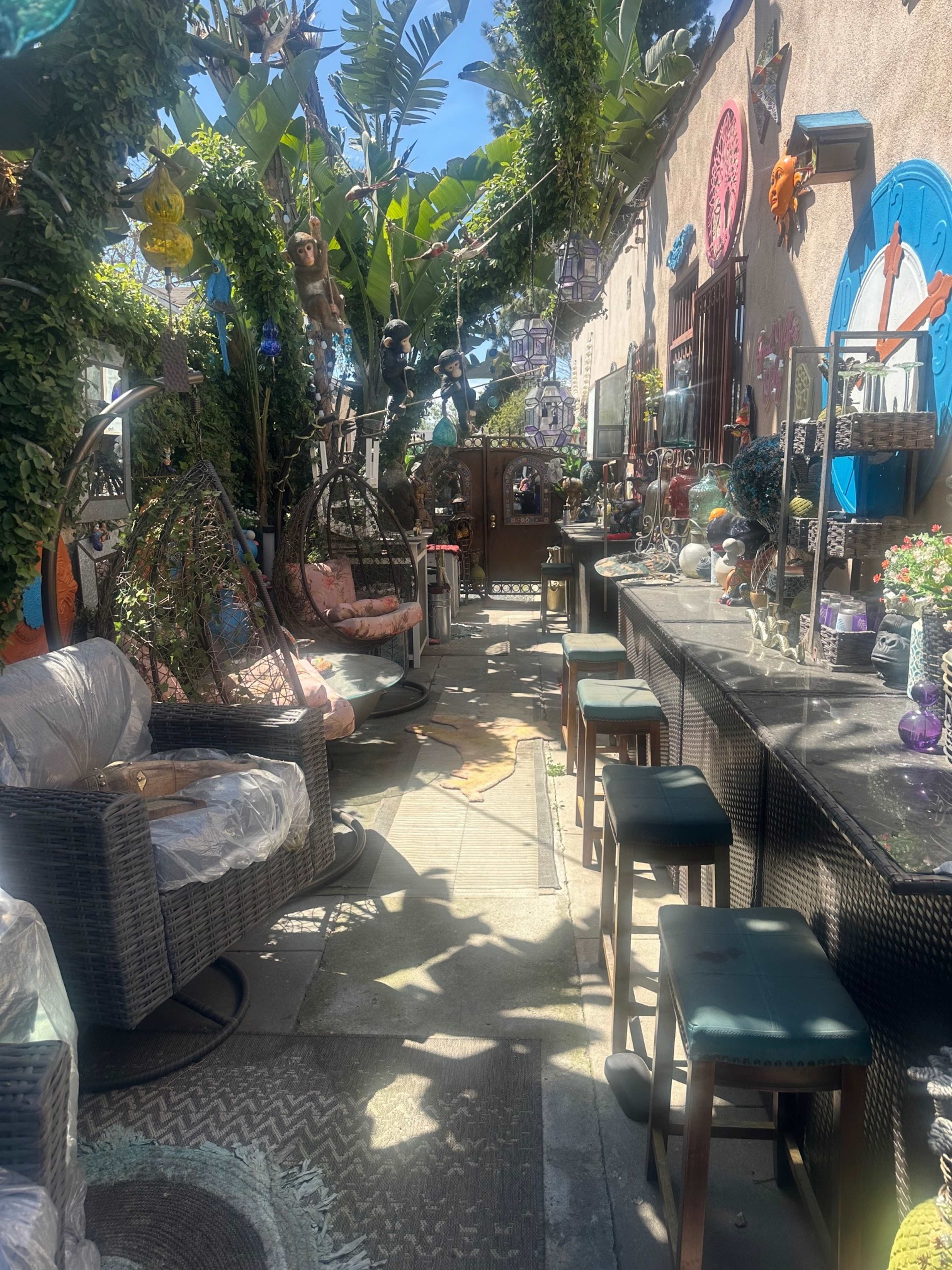 The image shows a colorful outdoor garden space featuring hanging chairs, bar stools, and various decorative items surrounded by lush greenery.