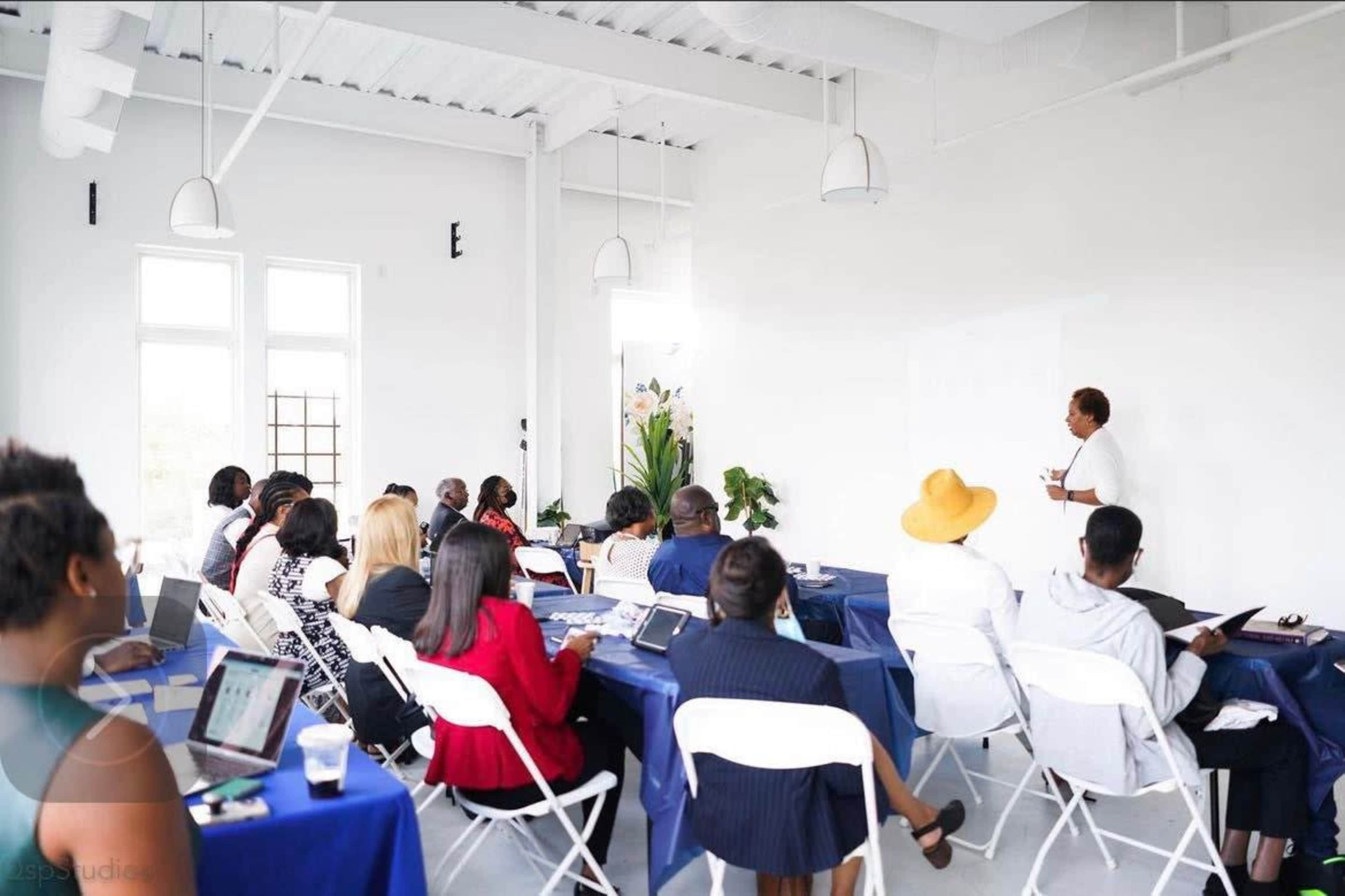 A group of people sits at tables in a bright room, facing a speaker who is presenting at the front.