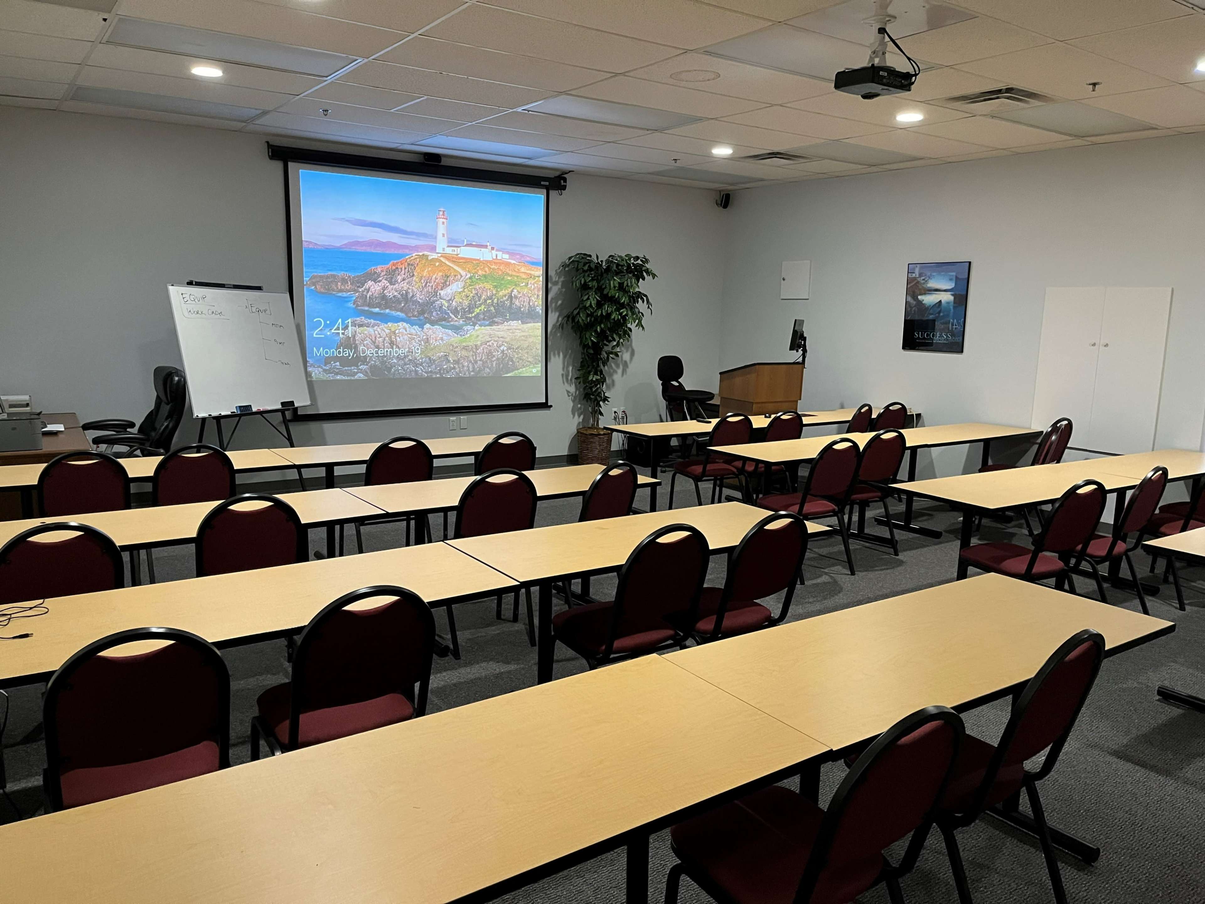 A conference room is set up with rows of tables and chairs, a projector screen displaying a lighthouse image, and a podium at the front.