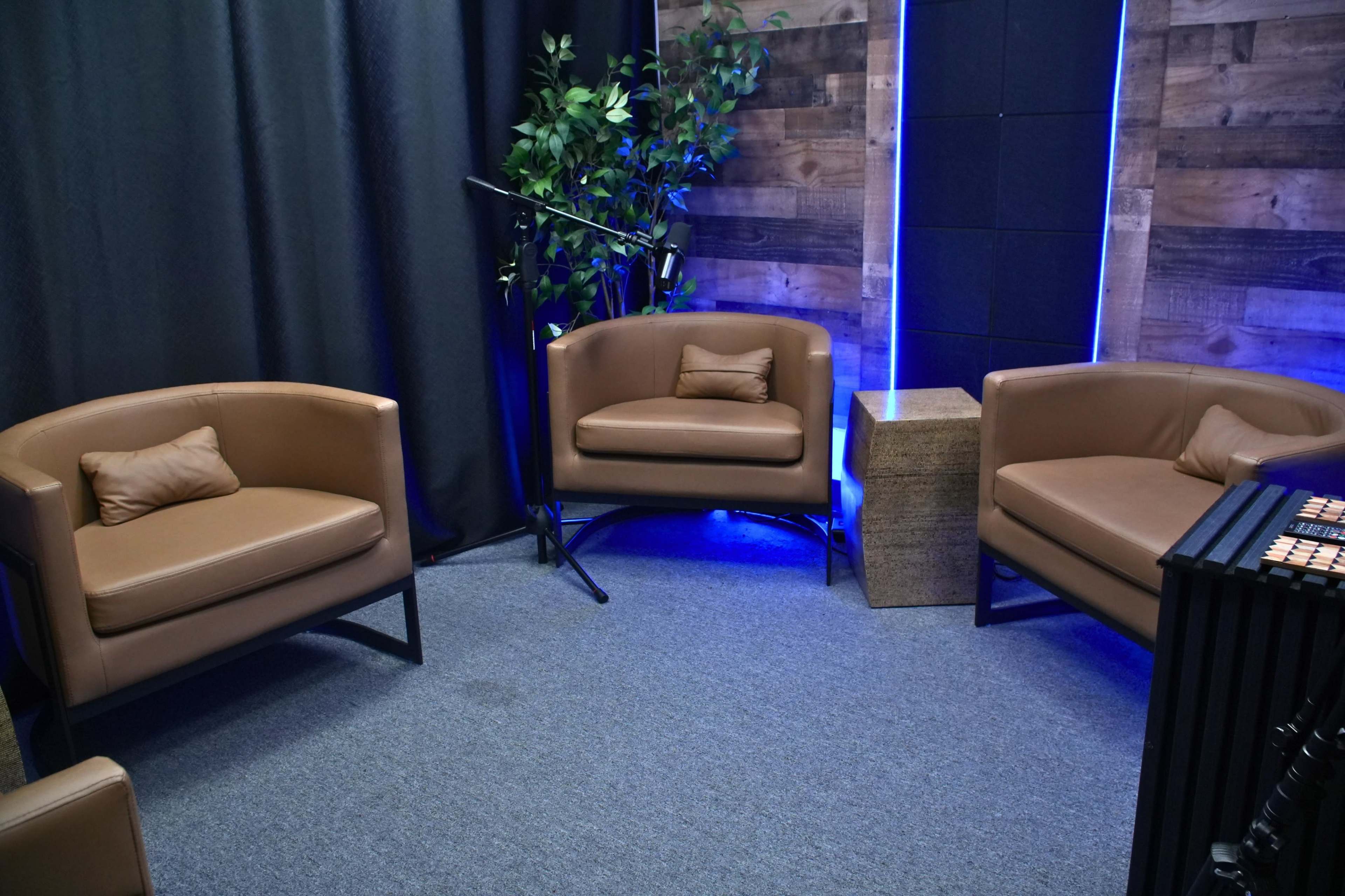 The image shows a small lounge area featuring two brown chairs, a small table, and a microphone setup against a backdrop of dark curtains and illuminated walls.