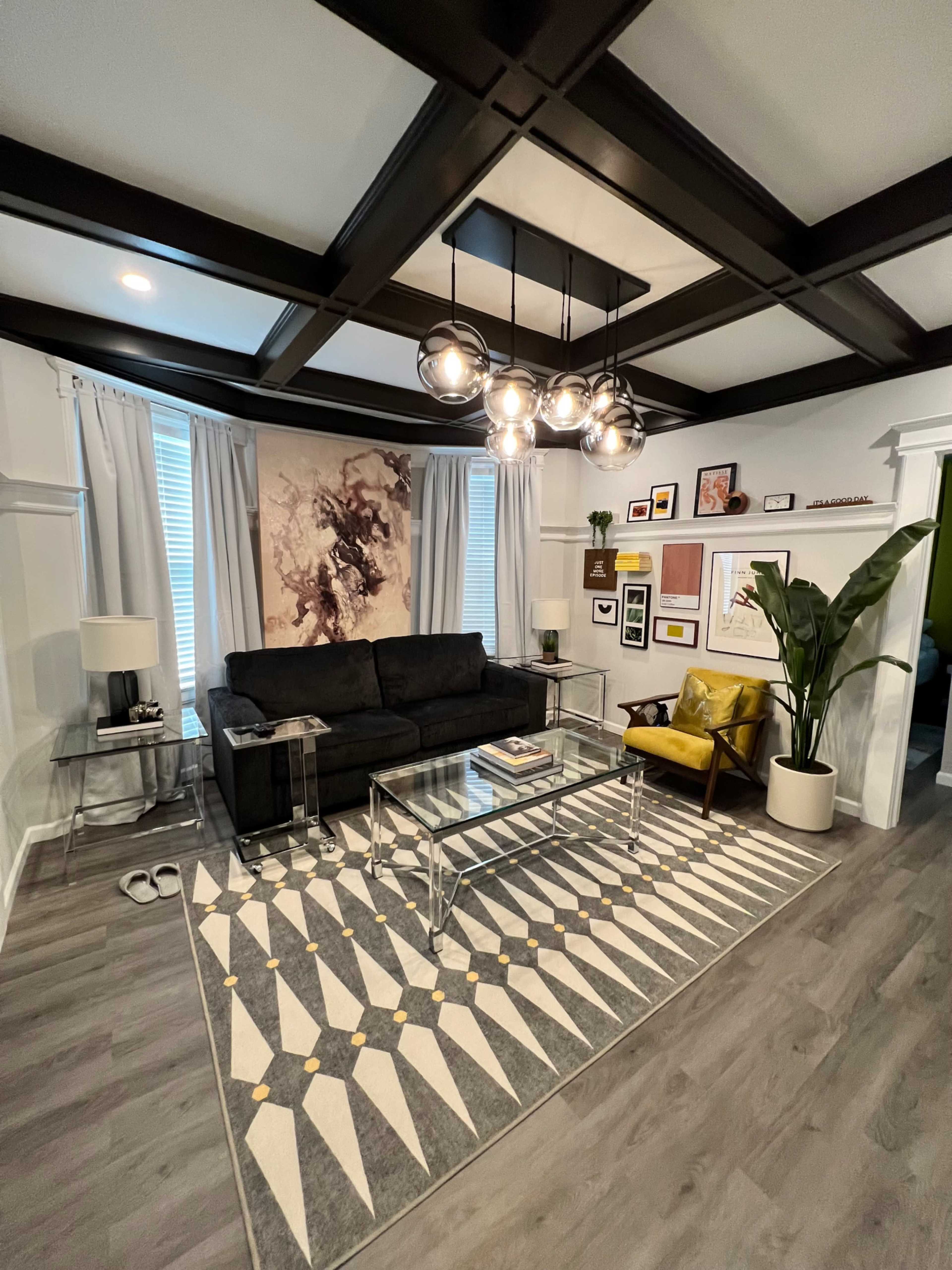 A modern living room featuring a black couch, a yellow armchair, a large wall art piece, and a coffee table on a patterned rug, with decorative plants and framed photographs on the walls.