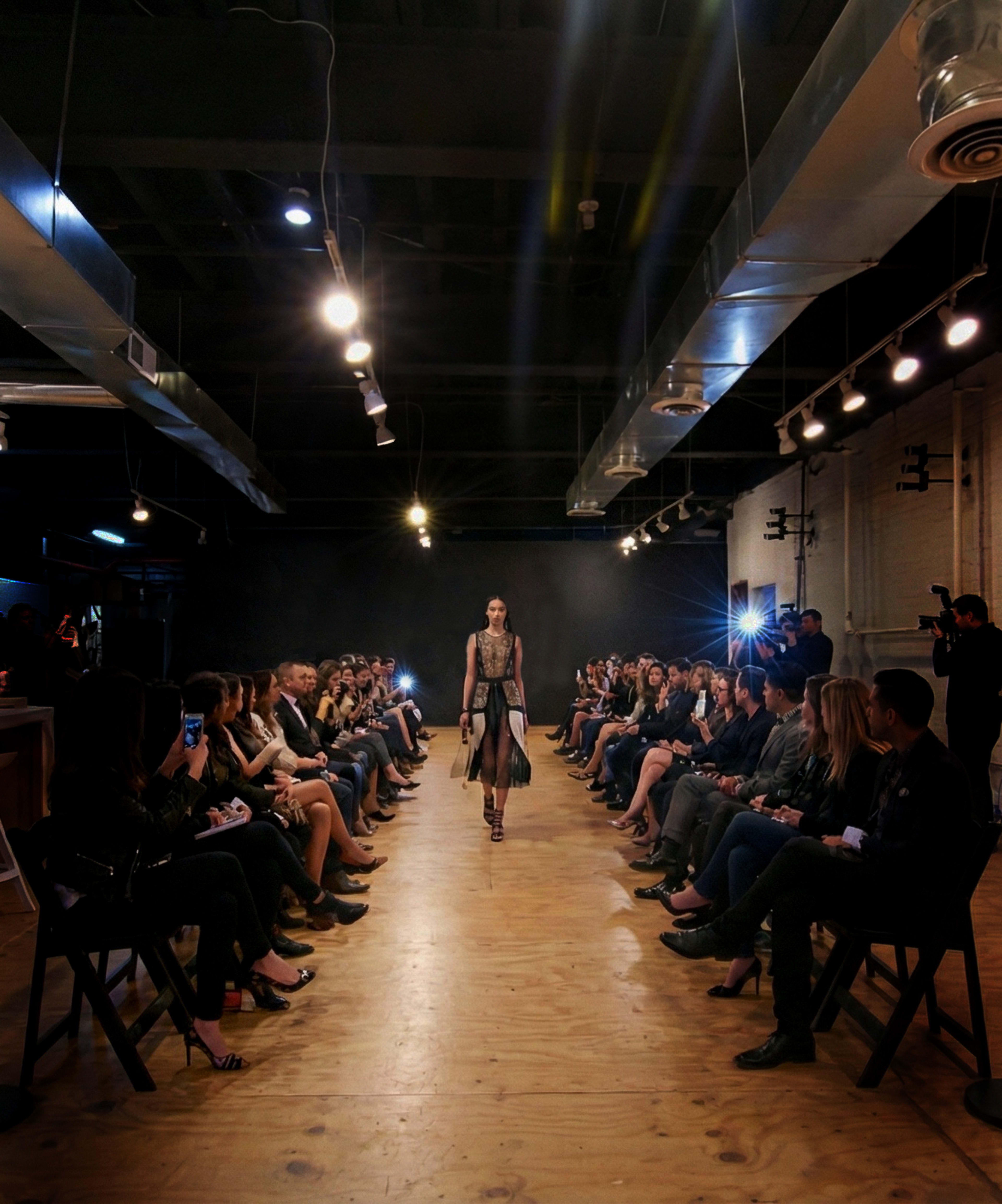 A model walks down a runway surrounded by an audience seated on both sides, with bright lights focused on the catwalk.