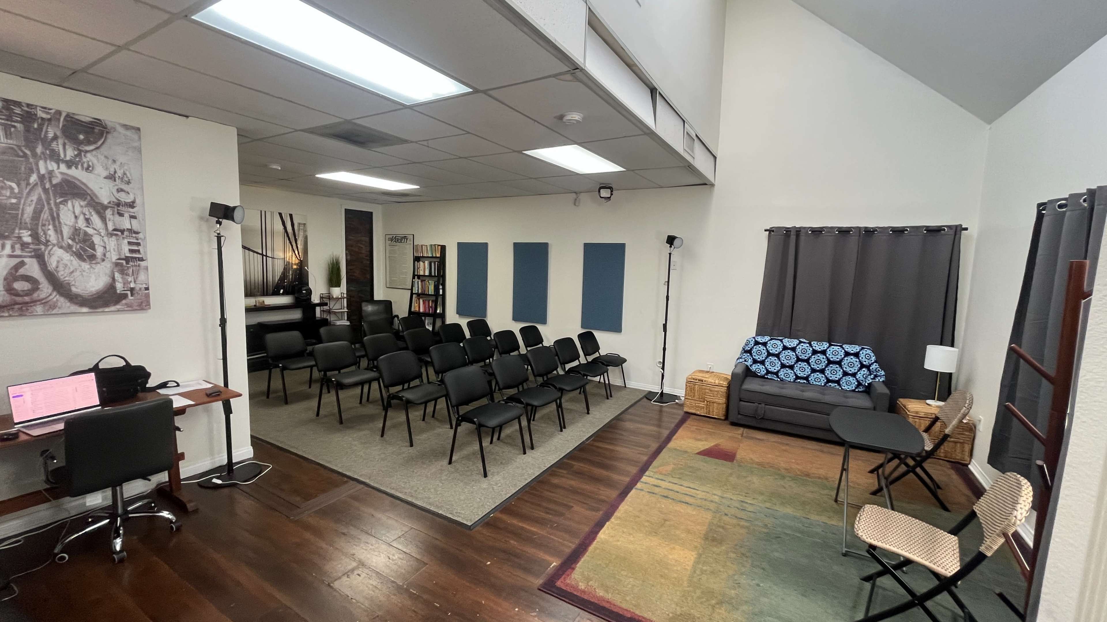 The image shows a multi-purpose room with a seating arrangement of black chairs facing a wall, a small table and chairs in the foreground, and artwork and shelves in the background.