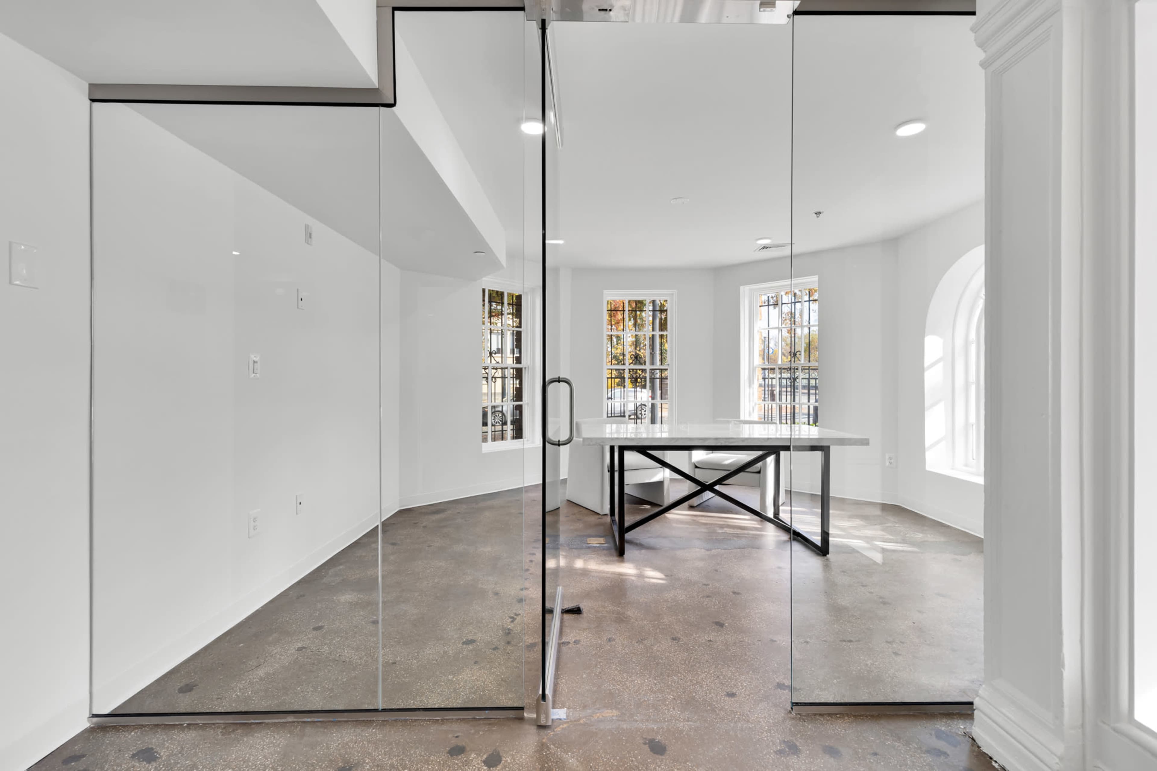 A glass door opens into a bright, minimalist office space featuring a large table and multiple windows.