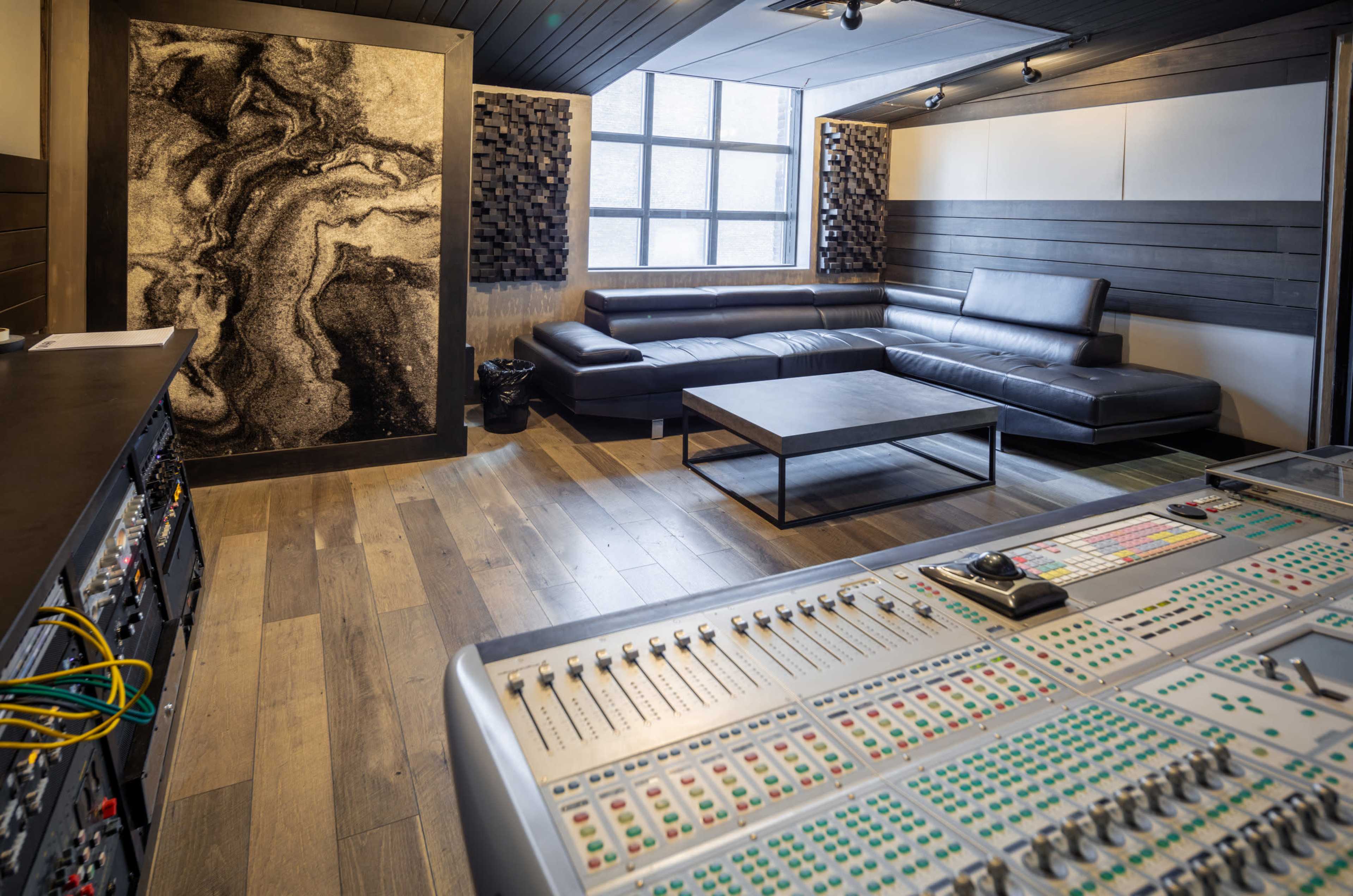 The image shows a modern recording studio featuring a control desk with multiple colorful knobs and sliders, a black leather sectional couch, and a large acoustic panel on the wall.