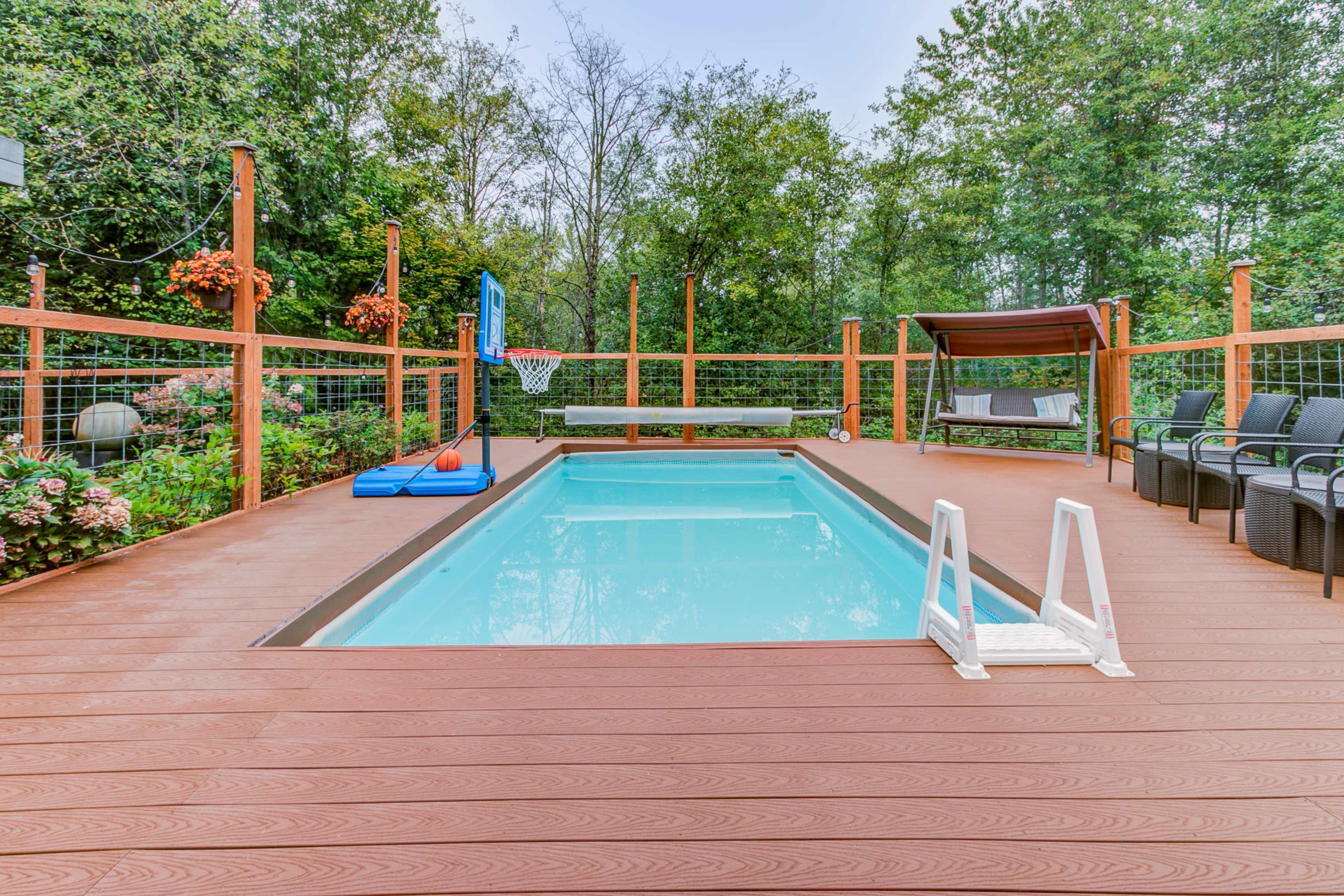 The image shows a swimming pool surrounded by a wooden deck, with a basketball hoop, seating area, and a shaded canopy nearby.