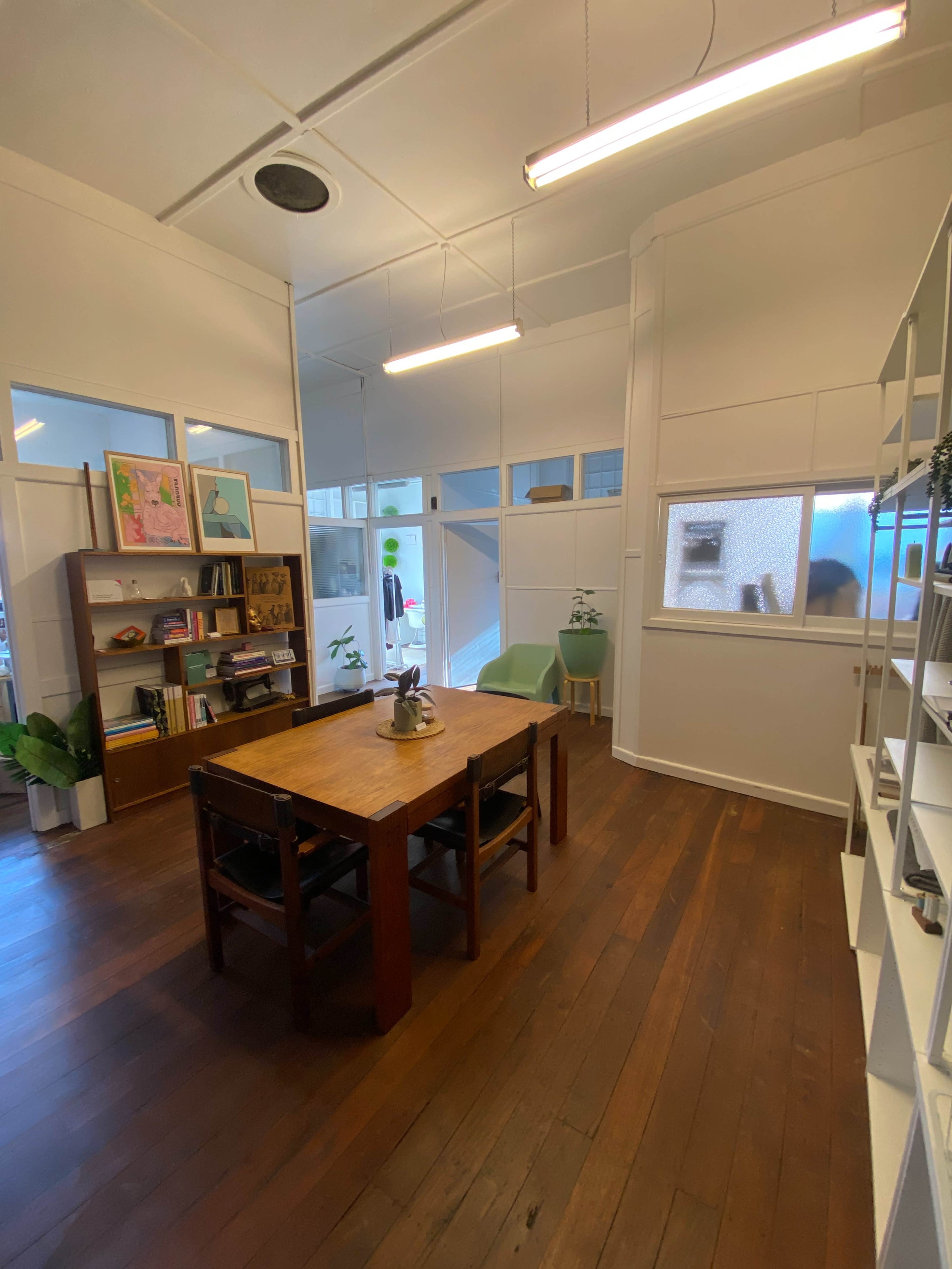 The image shows a bright room with wooden flooring, featuring a central table surrounded by chairs, a bookshelf filled with items, and large windows allowing natural light.
