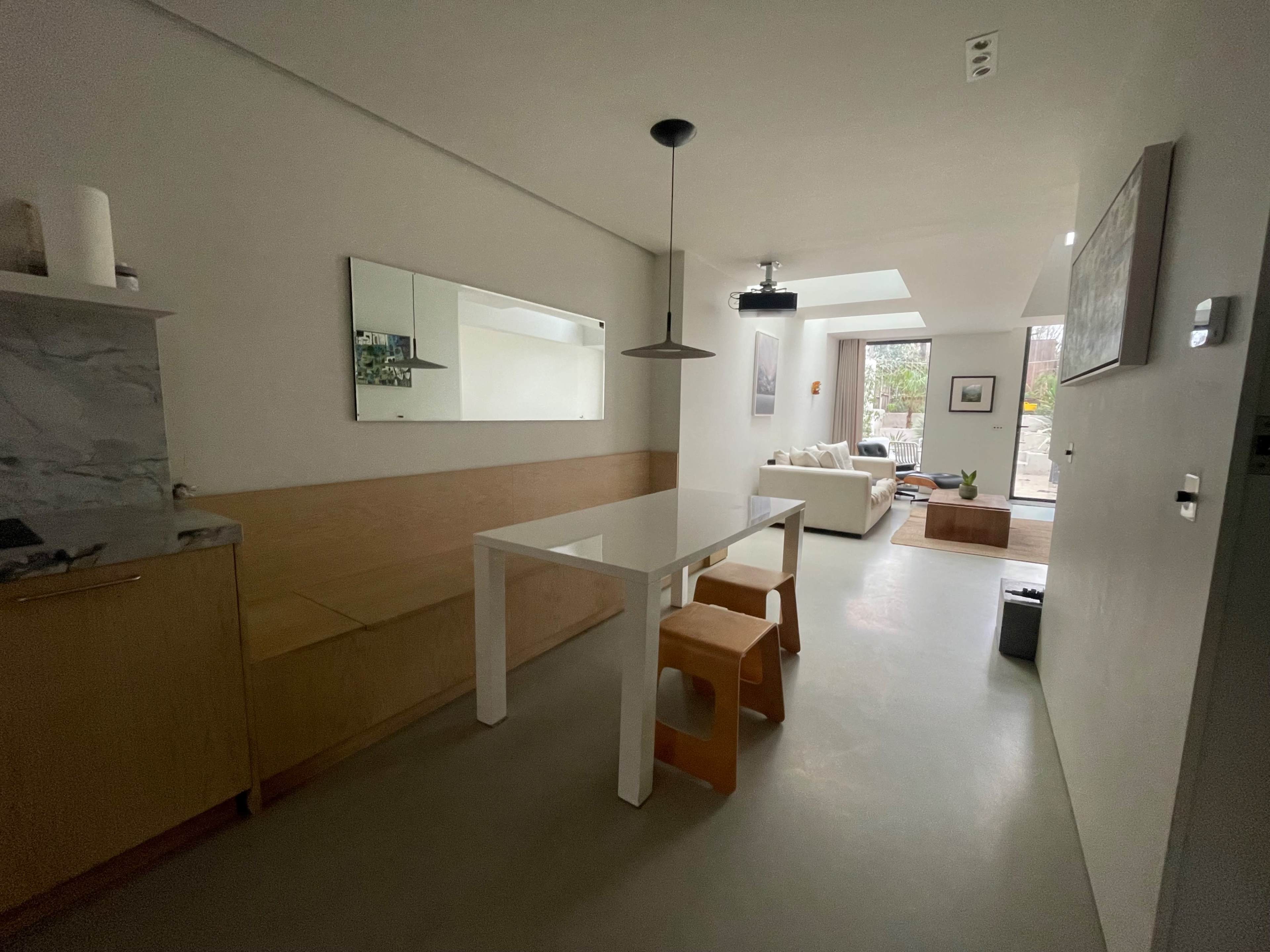 The image shows a modern, open-concept living space that includes a dining area with a white table and wooden benches, leading into a living room with a sofa and natural light coming through a skylight.