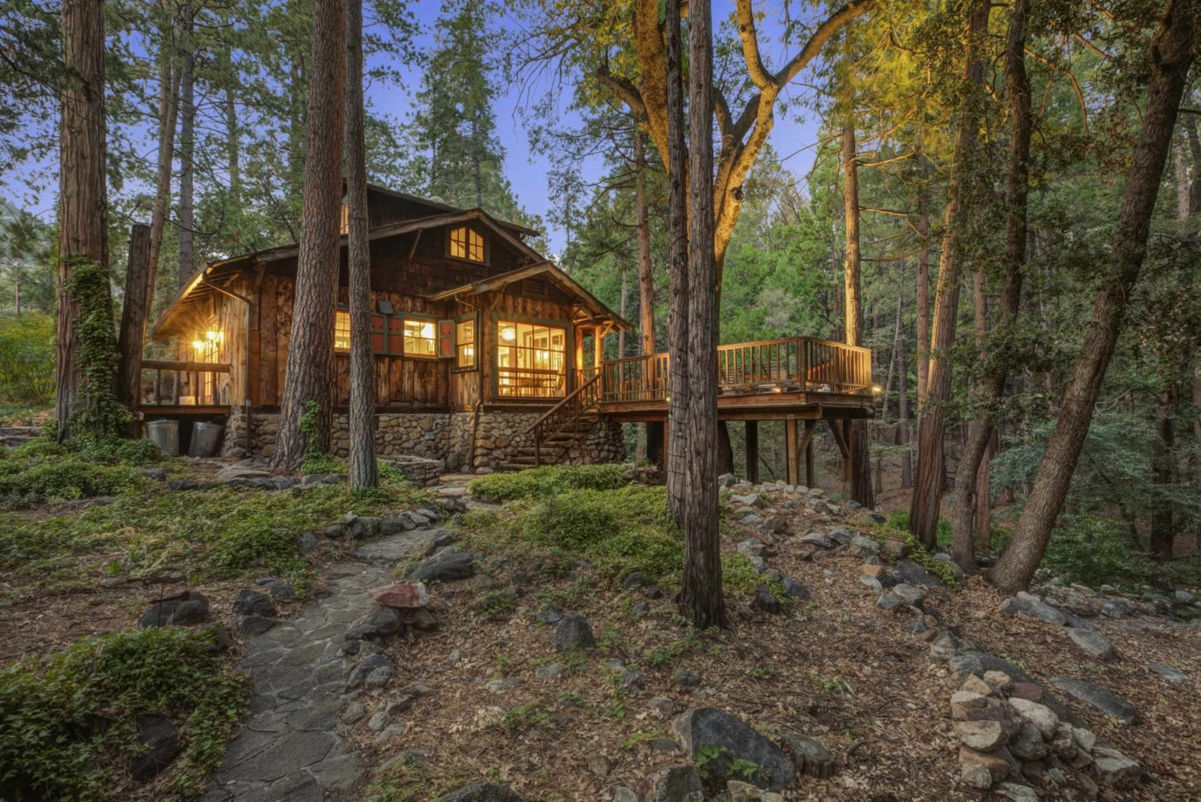 A rustic wooden cabin with large windows is situated among tall trees, surrounded by a rocky pathway and illuminated by warm indoor lights.
