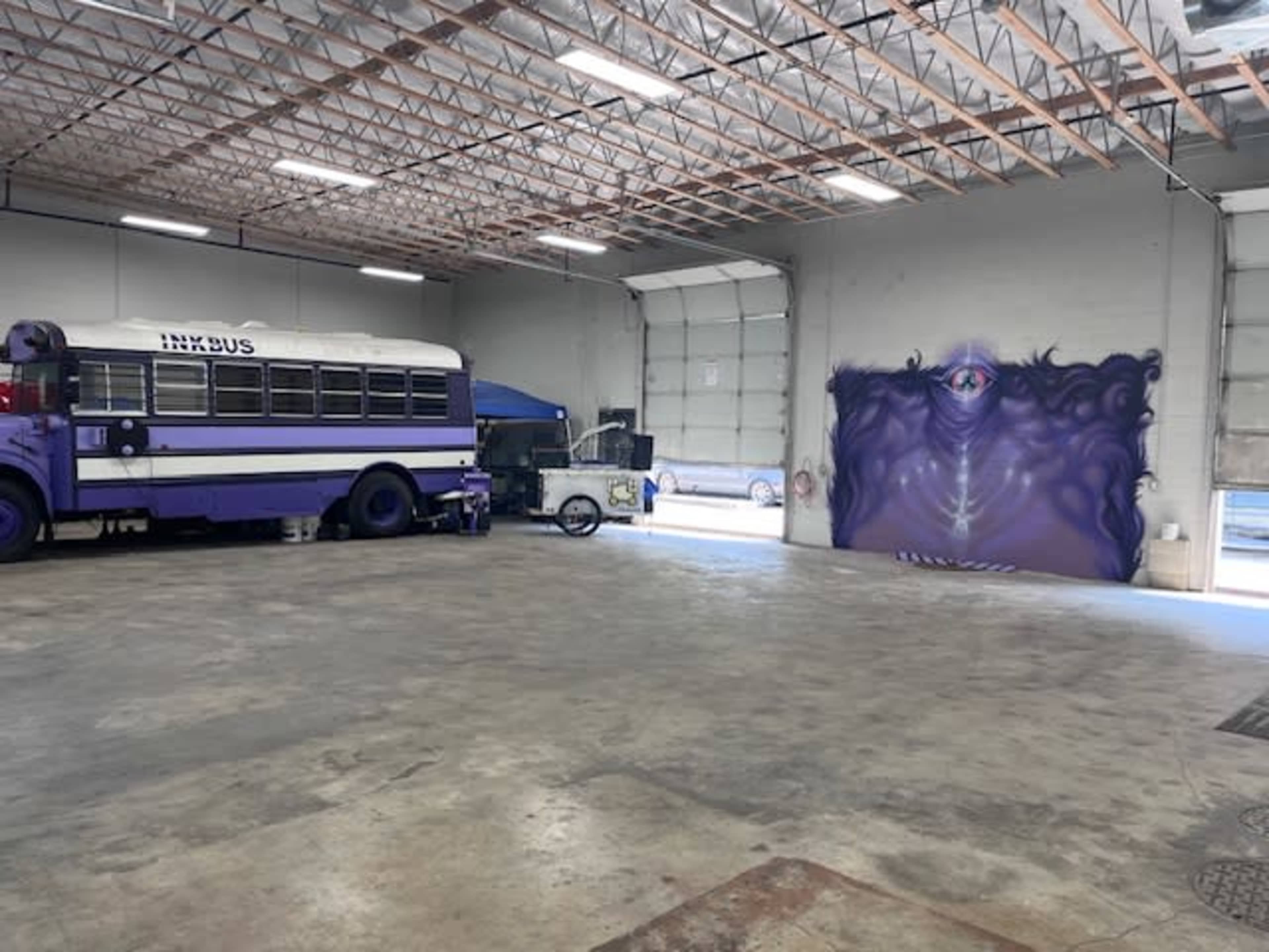 A purple school bus is parked inside a spacious industrial garage, with a mural of an eye on the gray wall.