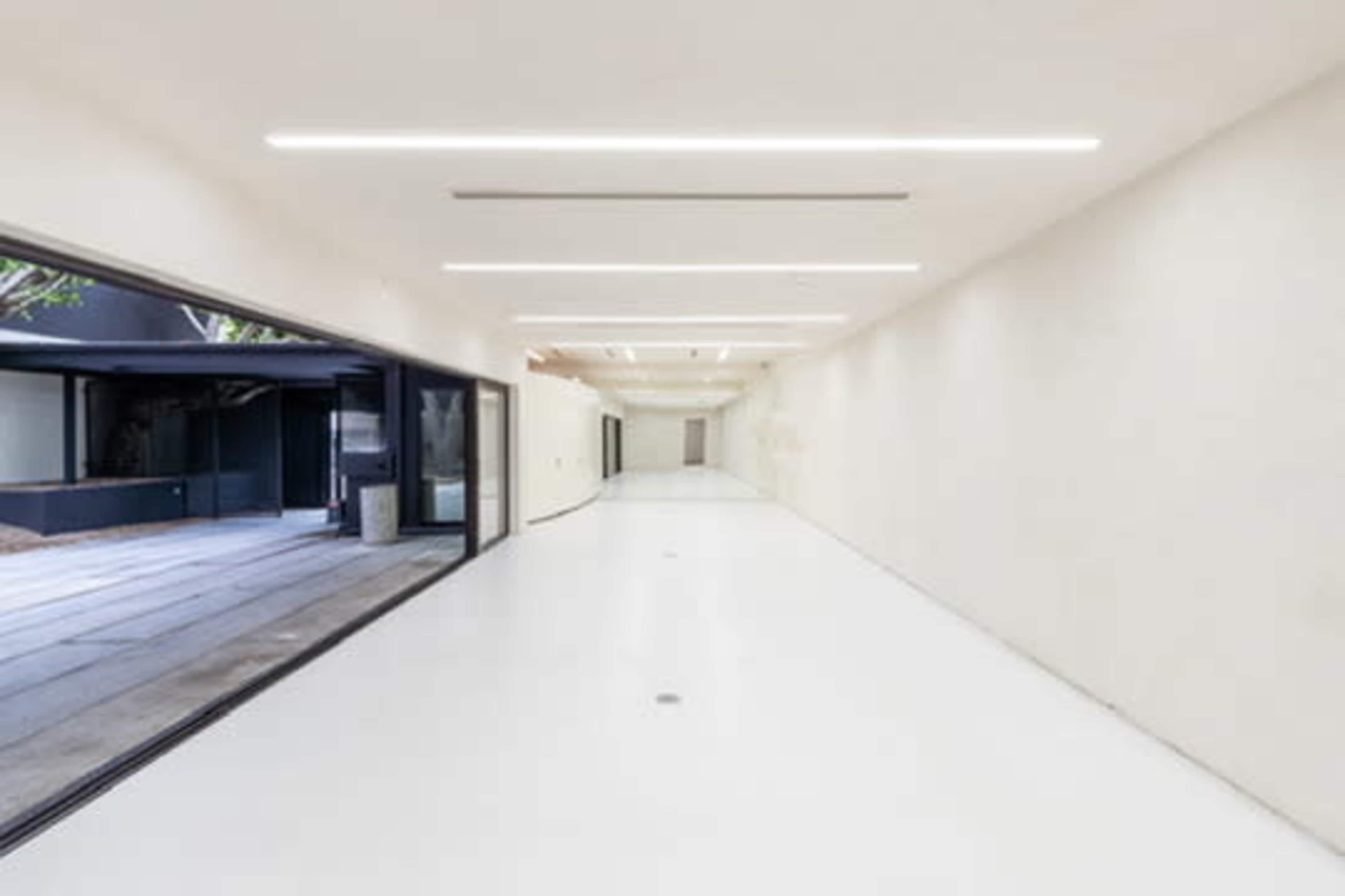 The image shows a spacious, minimalist hallway with white walls and flooring, illuminated by linear ceiling lights.