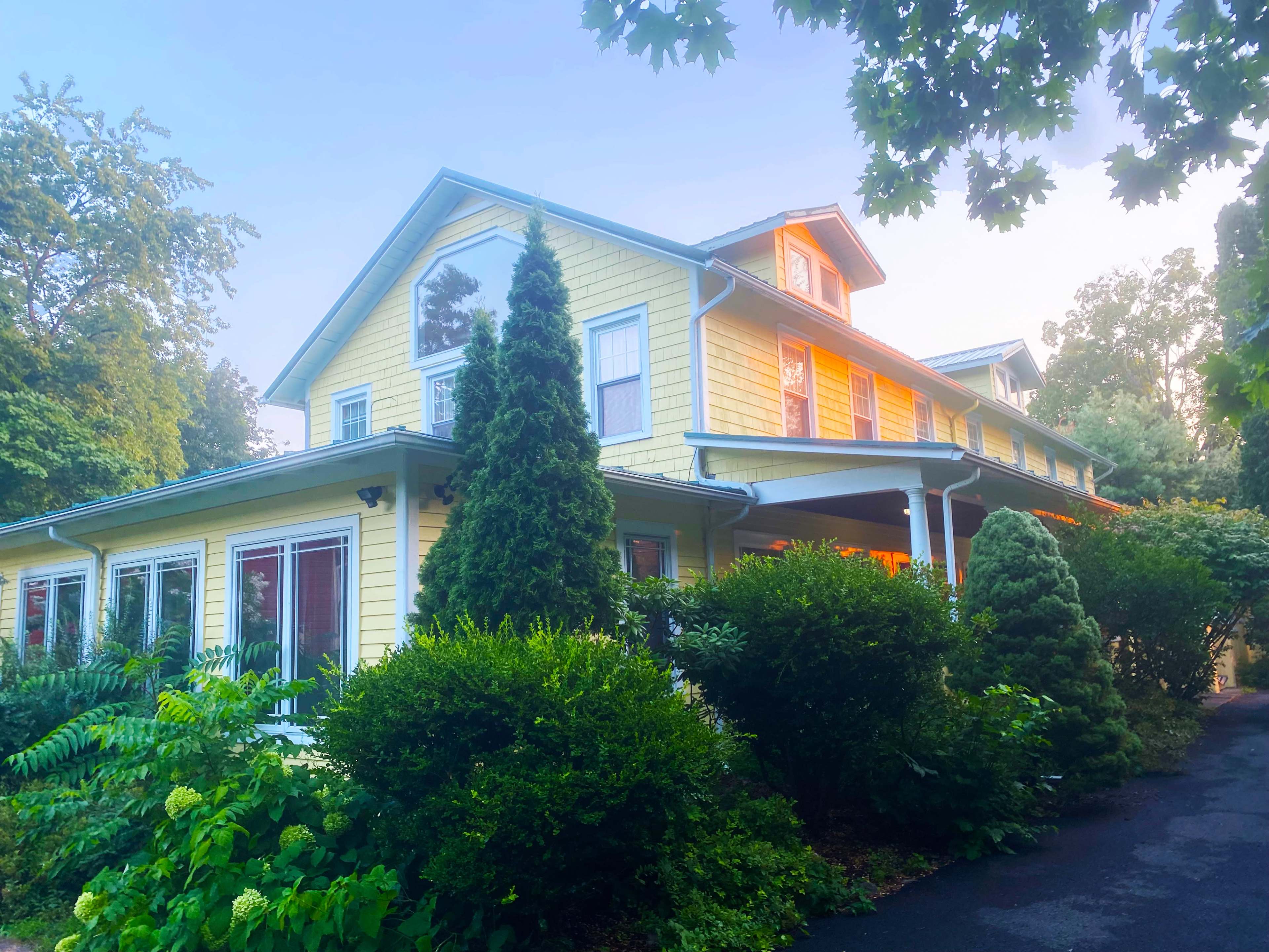 The image shows a large yellow house surrounded by green shrubs and trees.