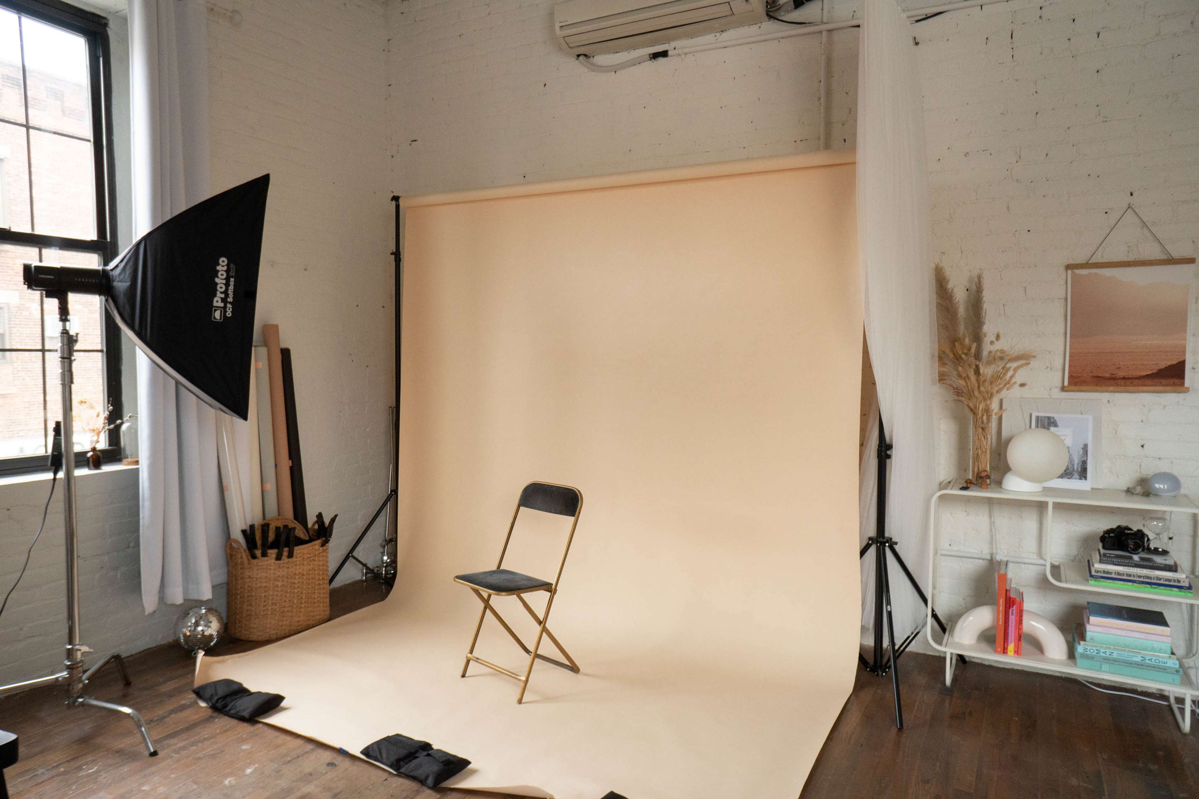 A photography setup features a empty folding chair in front of a neutral beige backdrop, with soft lighting equipment and decorative elements in a modern studio space.