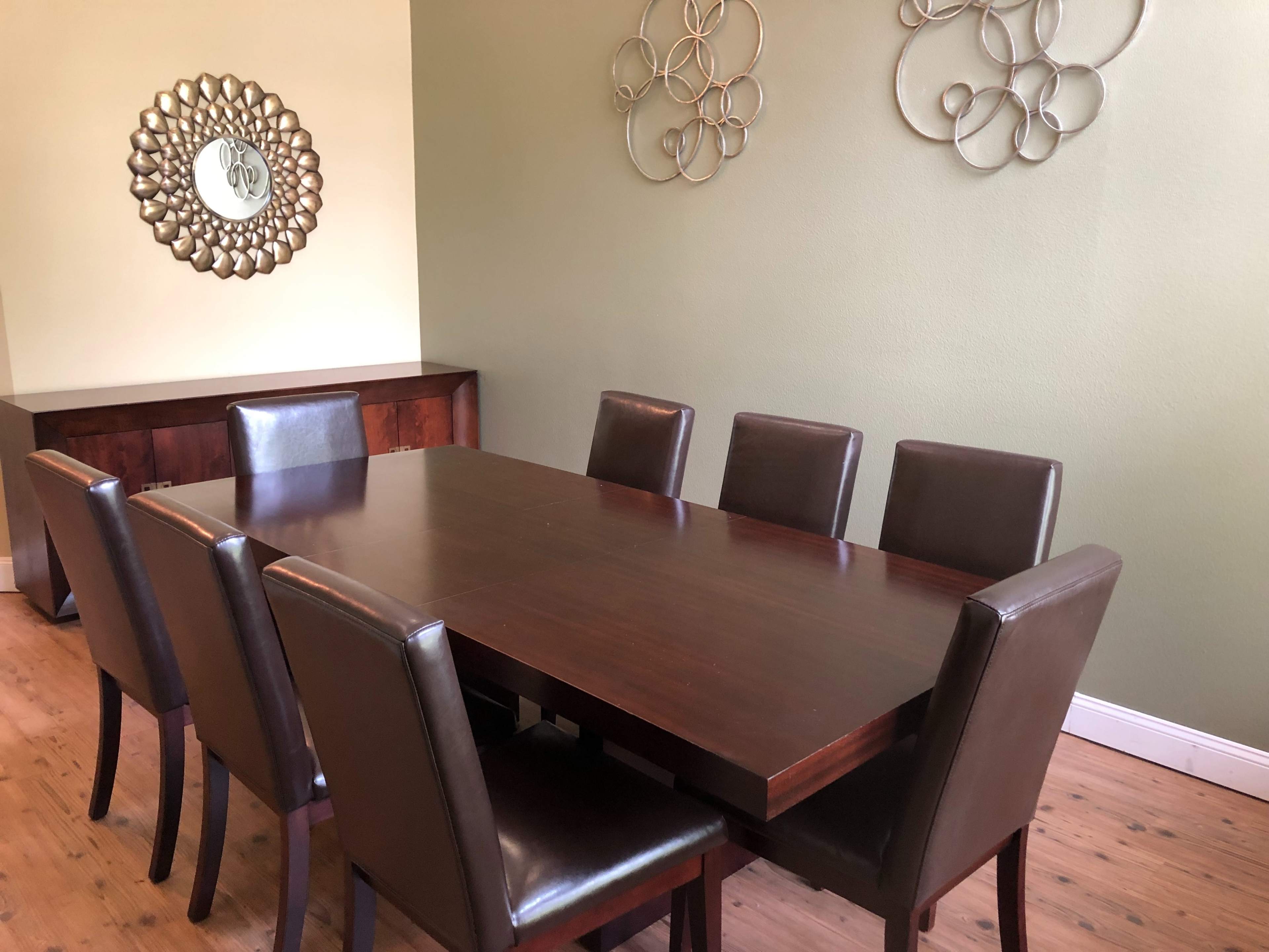 A wooden dining table with eight leather-upholstered chairs is positioned in a room with green walls and decorative metal wall art.