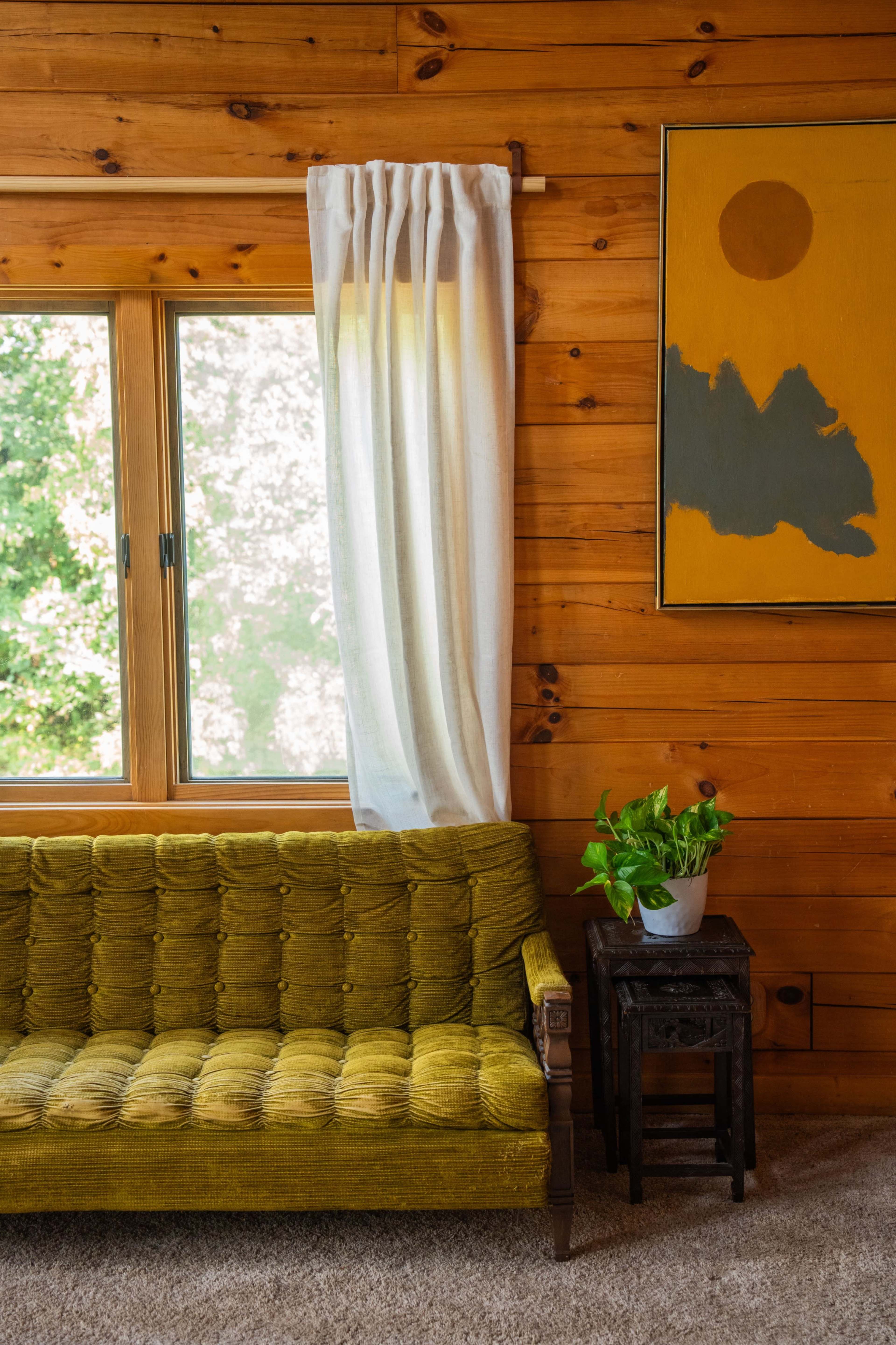 A cozy living room features a green tufted sofa next to a small table with a potted plant, set against a wooden wall and a large window.