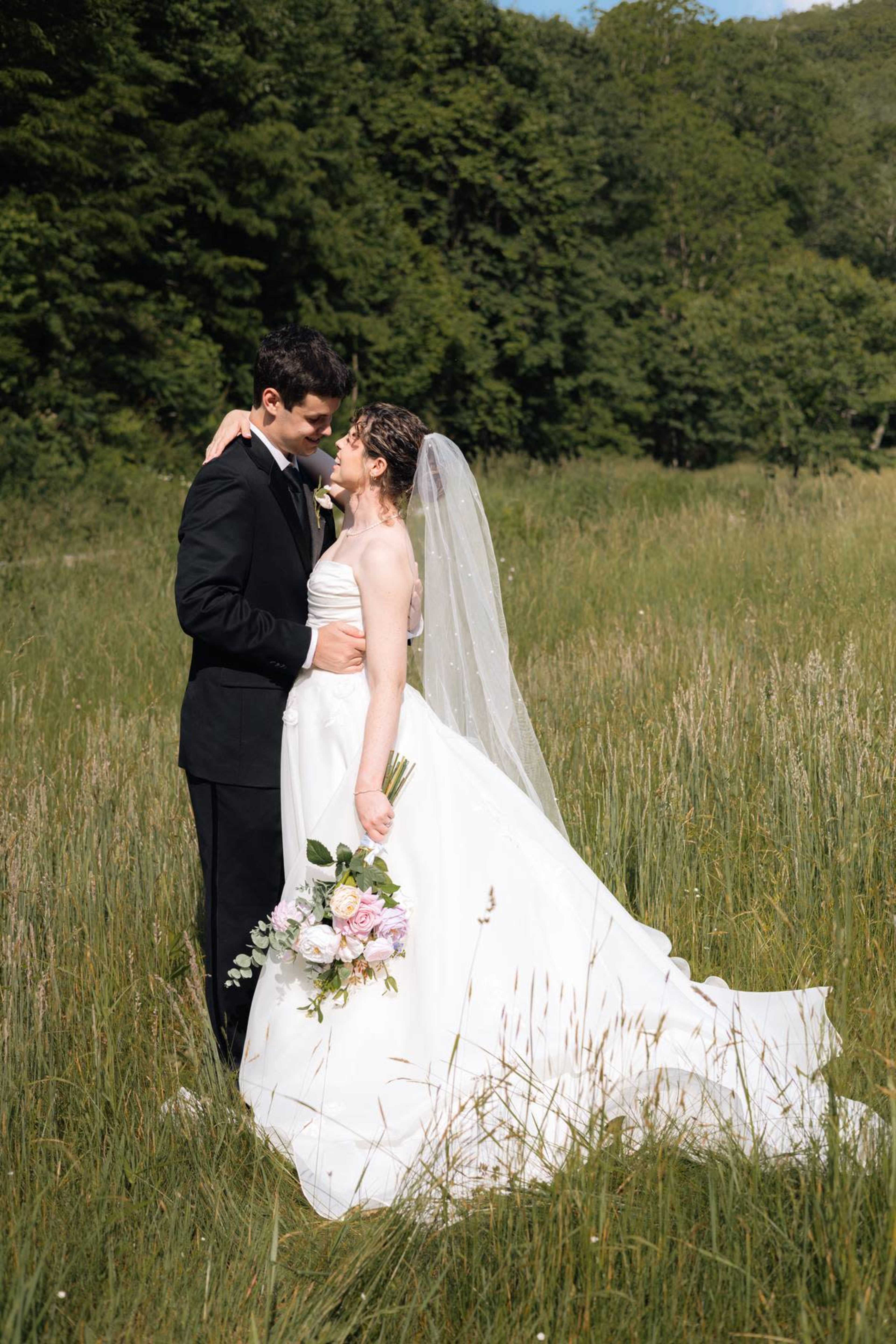 A couple shares a kiss in a grassy field, surrounded by trees, with the bride holding a bouquet of flowers.