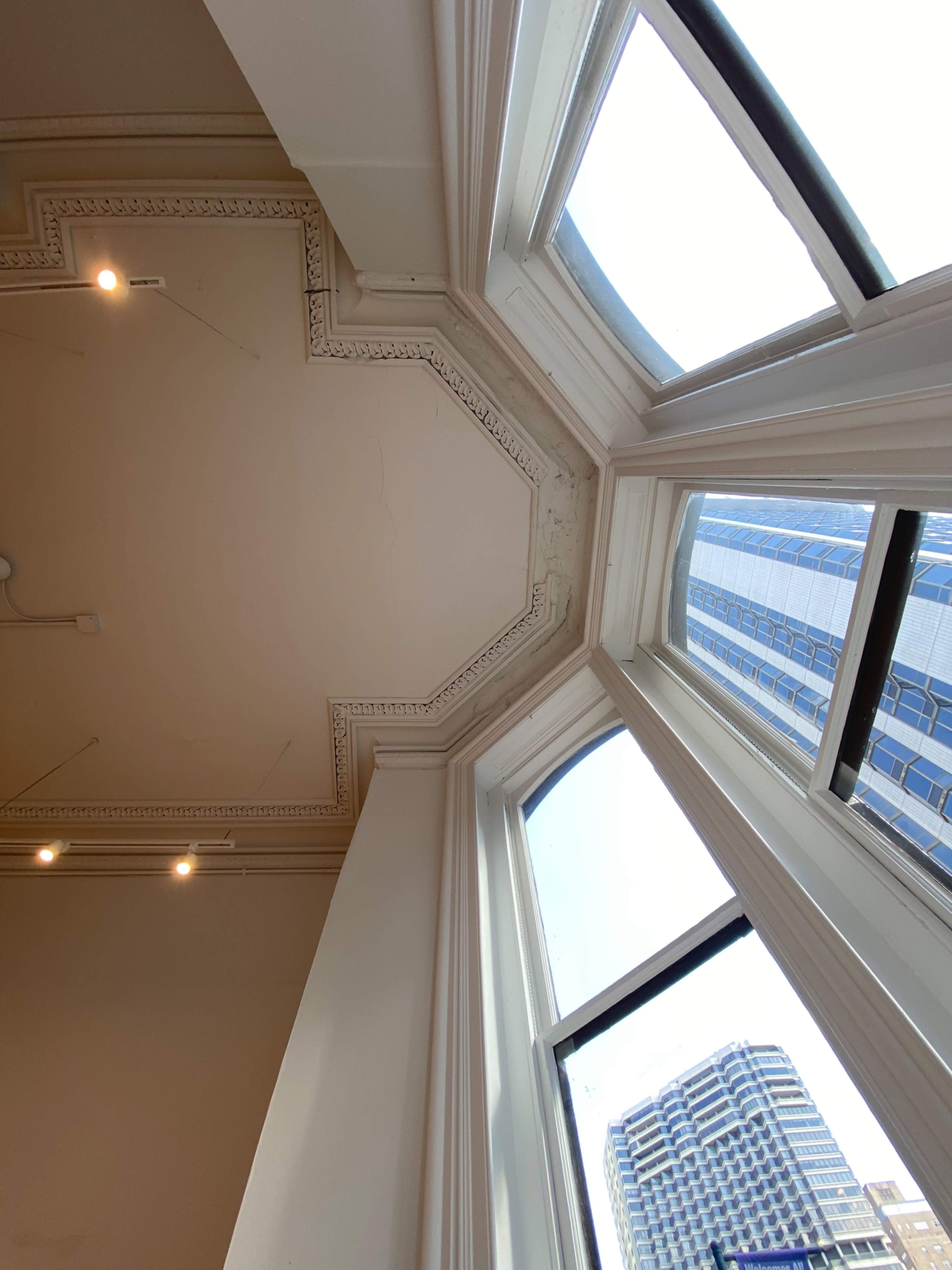 The image shows a view from the interior of a building, focusing on a decorated ceiling corner and large windows revealing a cityscape.