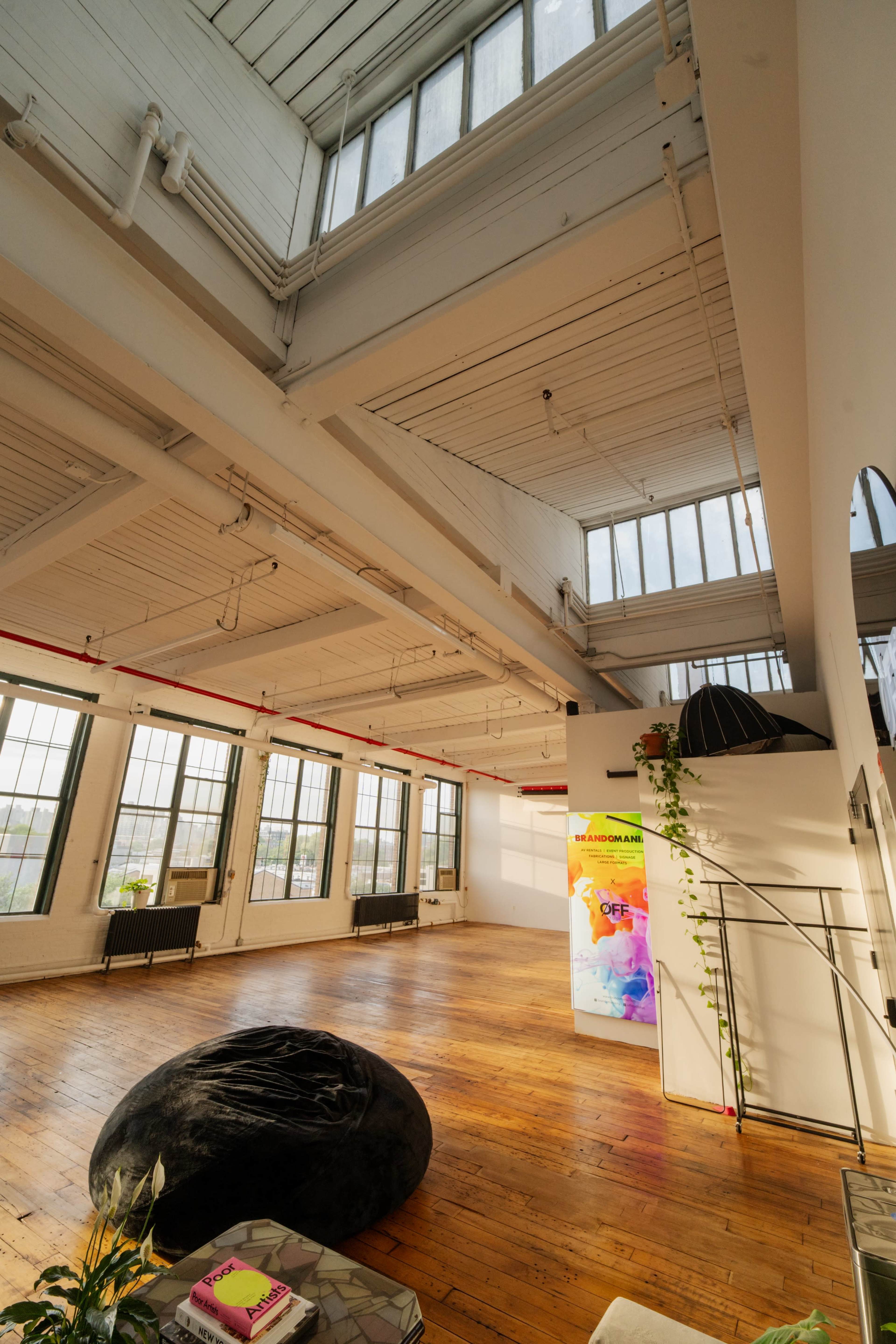 The image shows a bright, open space with large windows, hardwood floors, and minimalist furnishings.