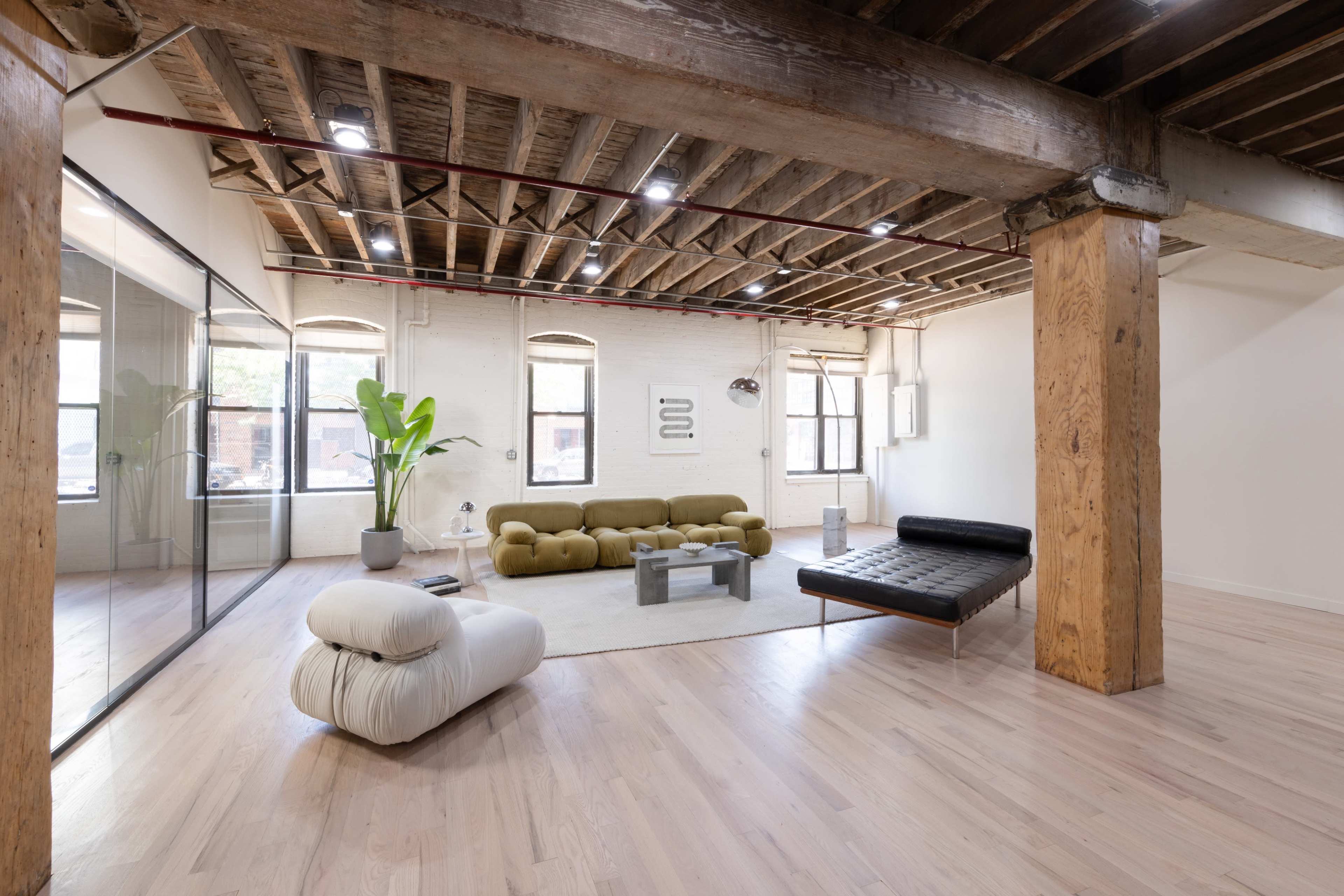 The image shows a modern, spacious living area with large windows, exposed wooden beams, and contemporary furniture including a green sofa and a black lounge chair.