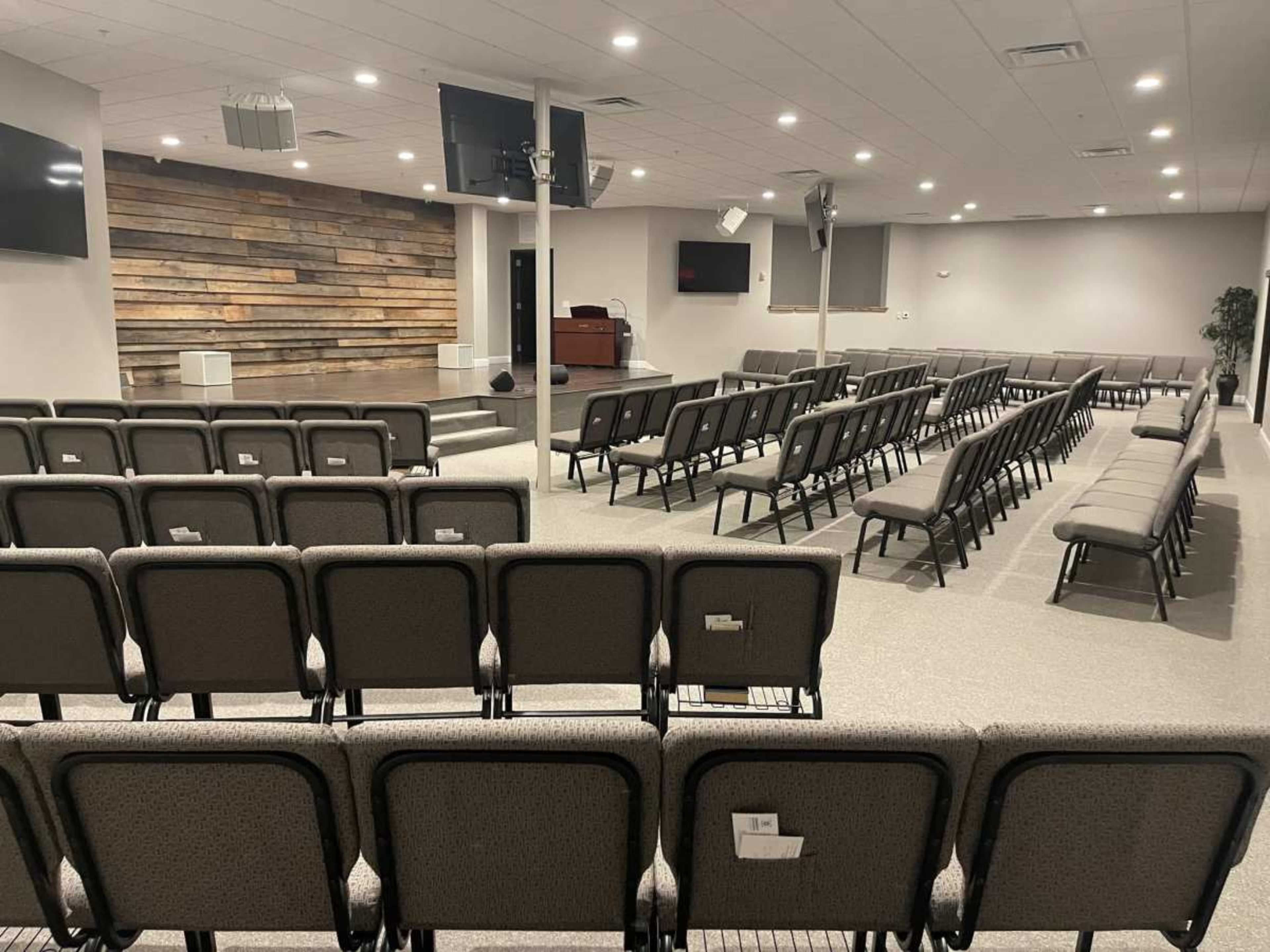 The image shows an empty indoor auditorium with rows of chairs facing a wooden-panelled stage area.